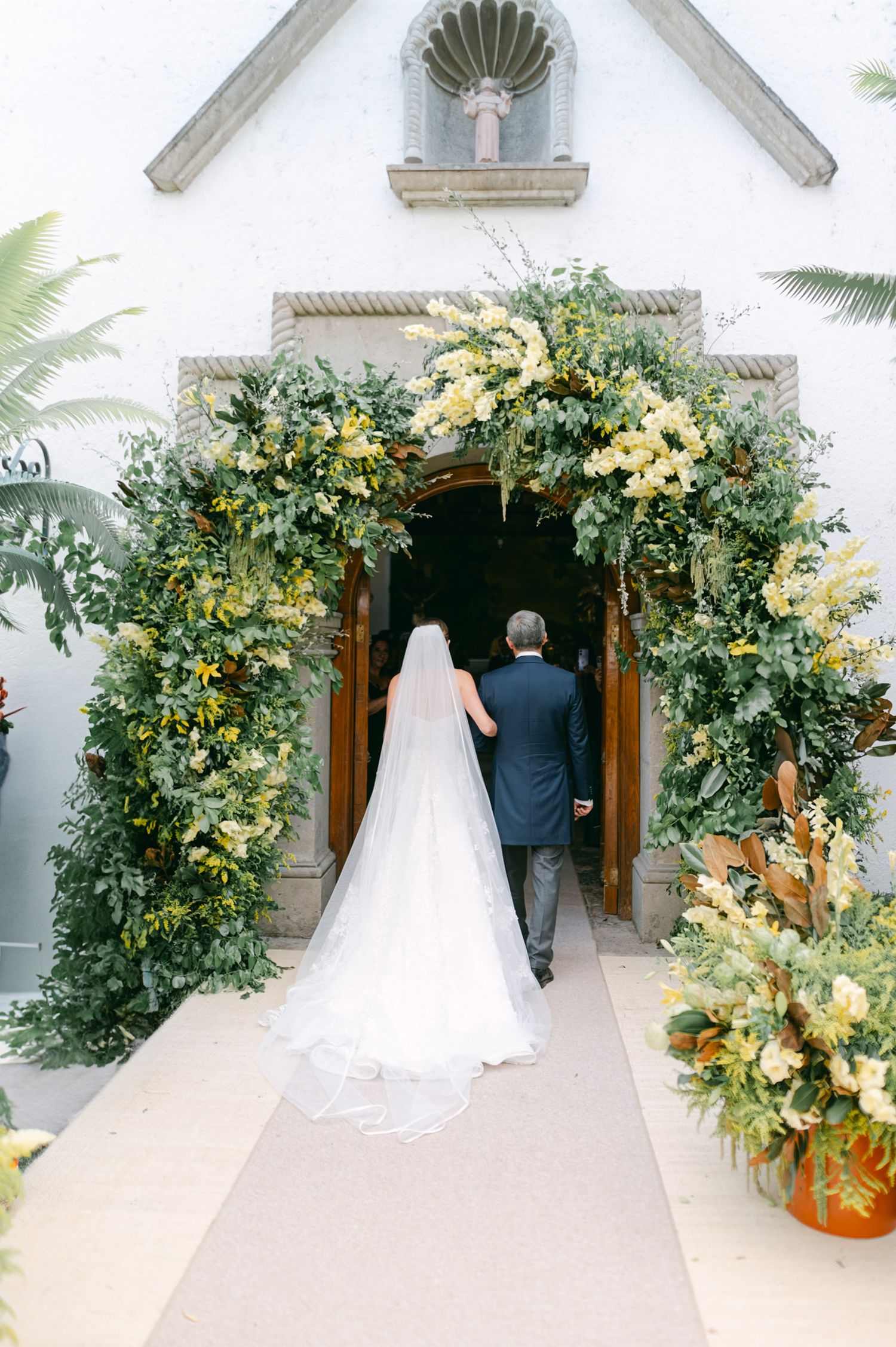 White chapel entrance adorned with lush greenery and yellow flowers for a wedding ceremony.