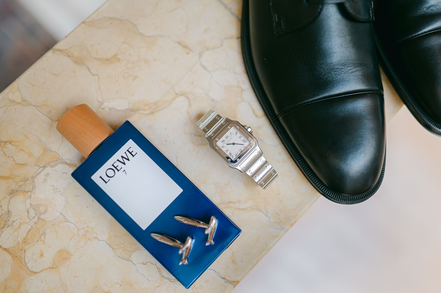 Groom wedding details flat lay featuring Loewe cologne, Cartier watch, cufflinks, and black dress shoes.