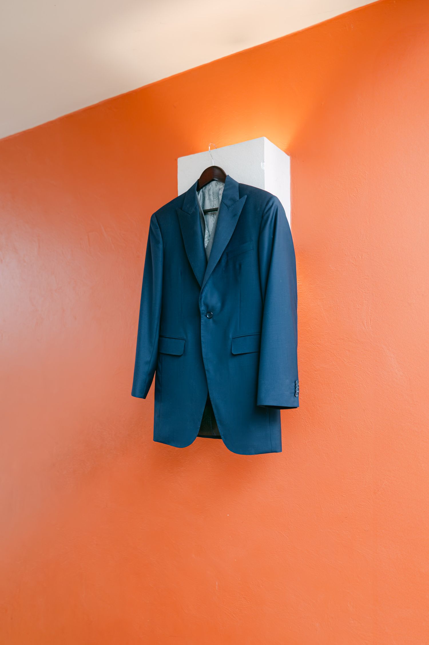 A blue teal suit jacket hanging on a wooden hanger against a bright orange wall.