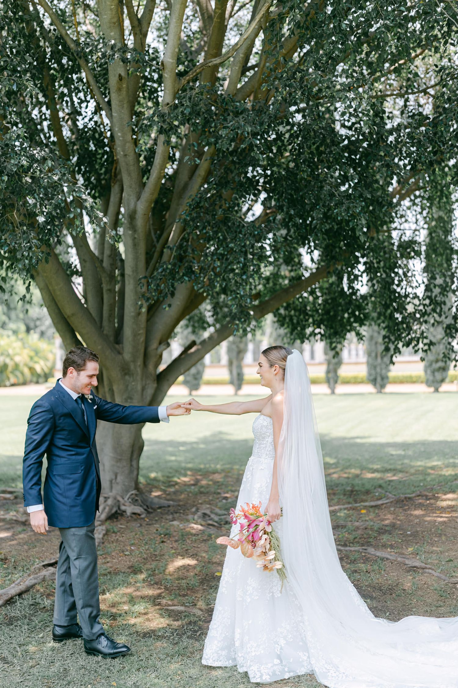 Groom spinning bride in lace wedding dress and cathedral veil under a large tree outdoors.