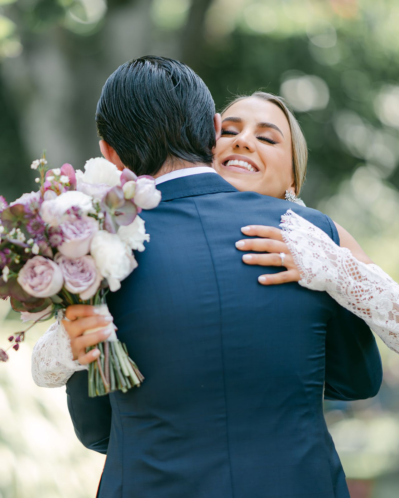 Smiling bride in lace gown embracing groom in navy suit while holding a floral bouquet.