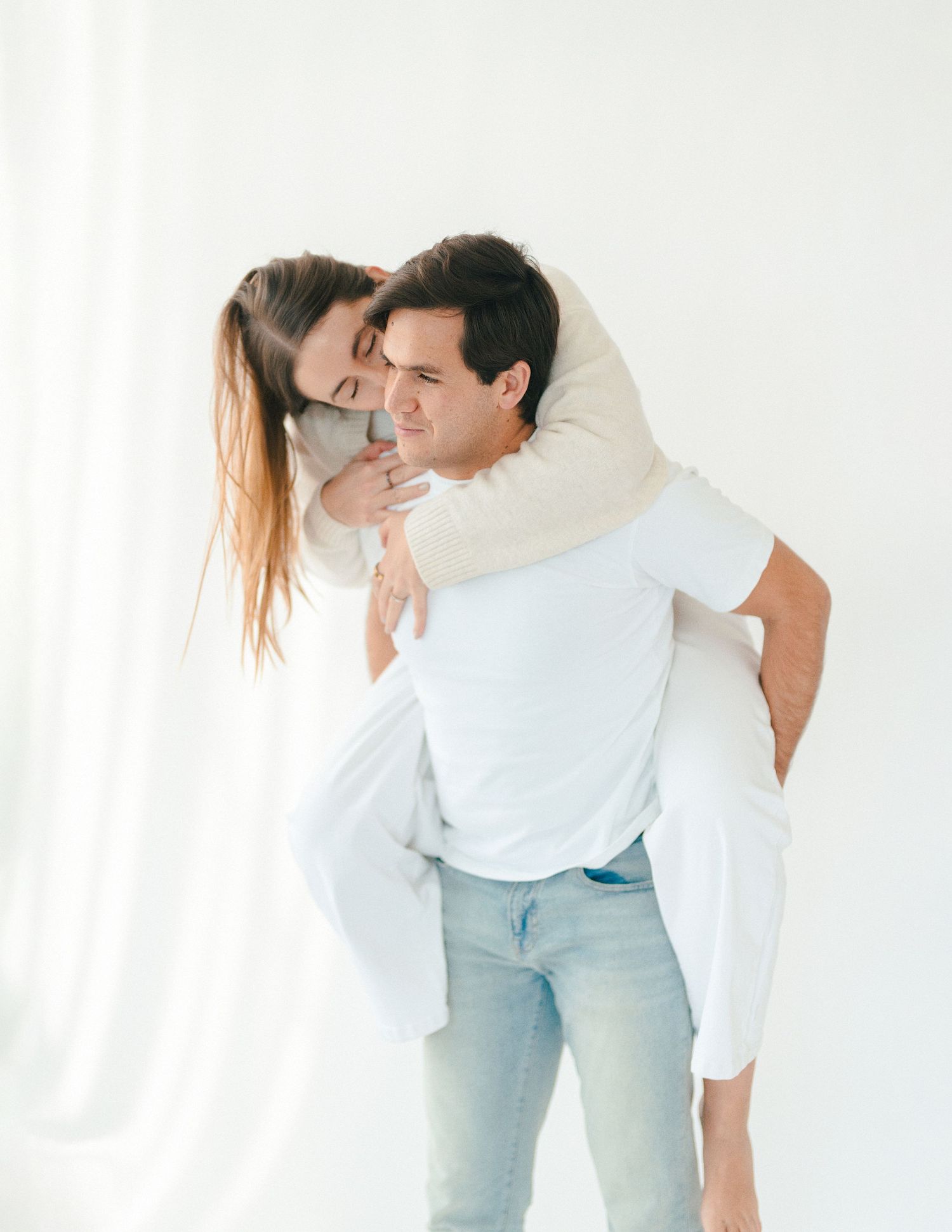 A laughing couple sharing a playful piggyback ride moment in a softly lit white studio backdrop.