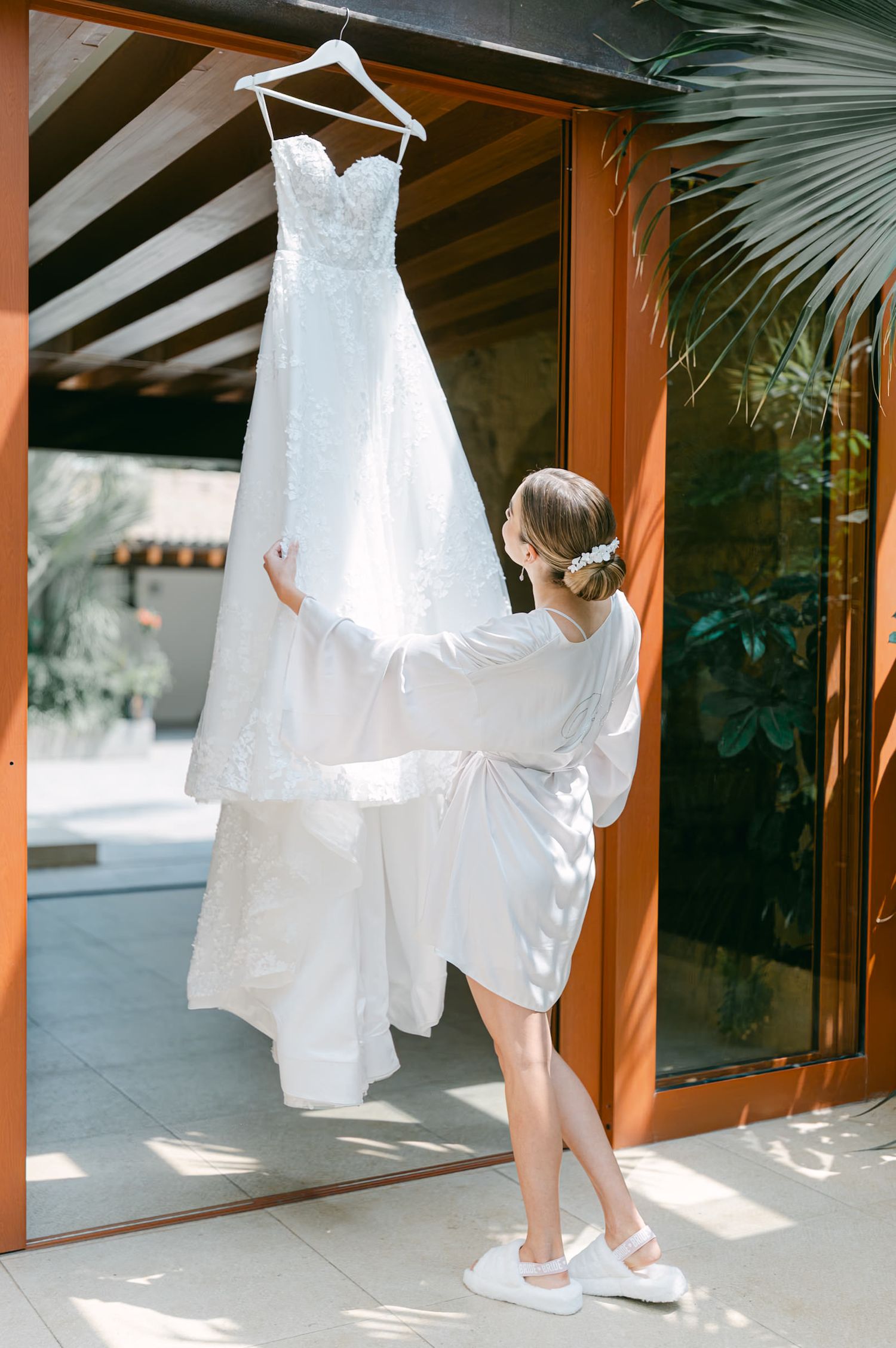 A bride in a white robe reaches up to touch her lace wedding gown hanging outdoors.