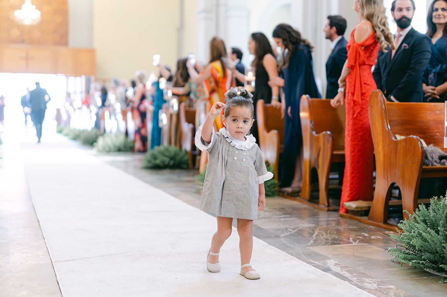 Young flower girl in gray dress walks alone down a sunlit church wedding aisle.
