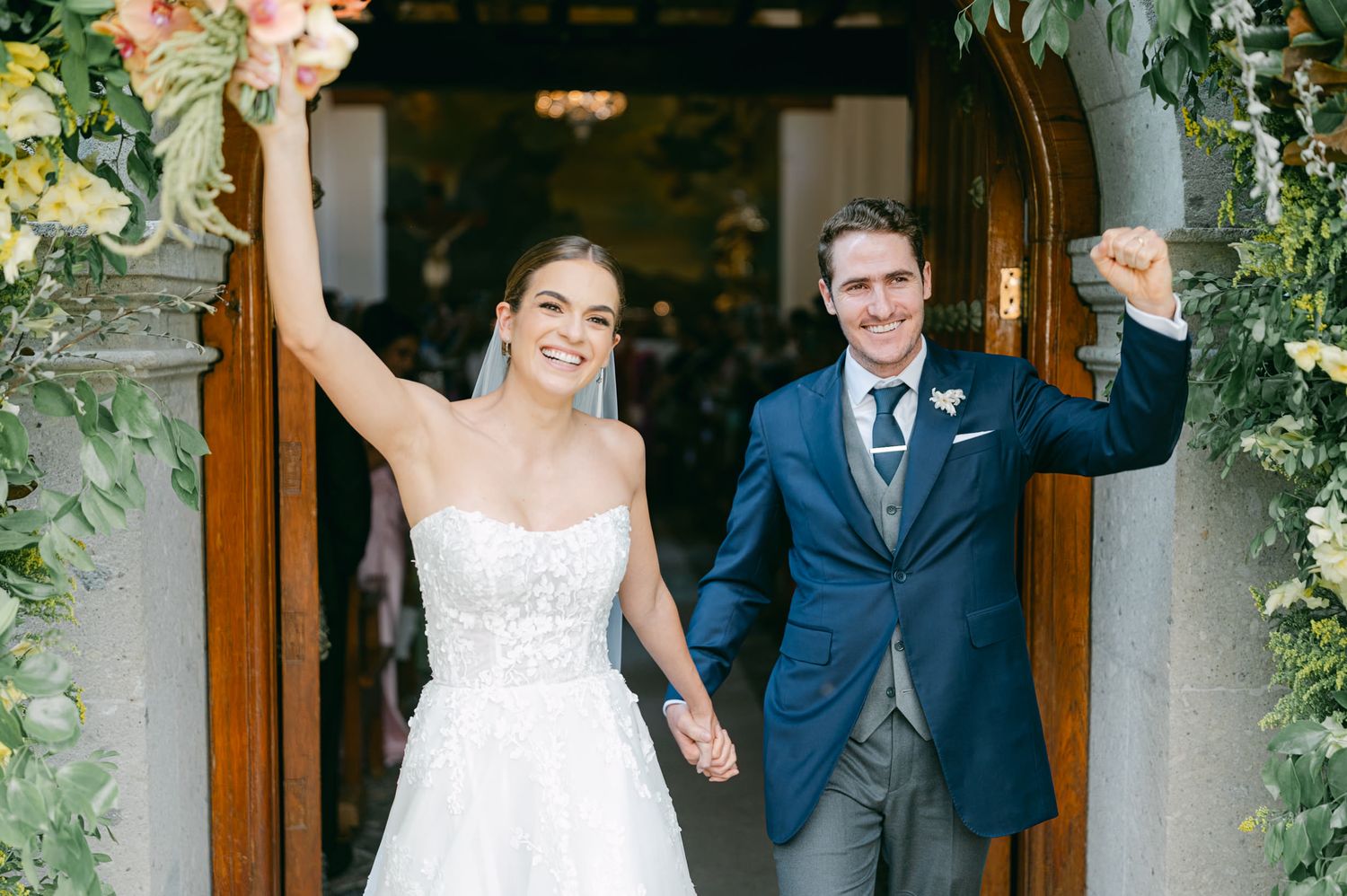 Couple exits through wooden church doors decorated with lush greenery and floral arrangements.