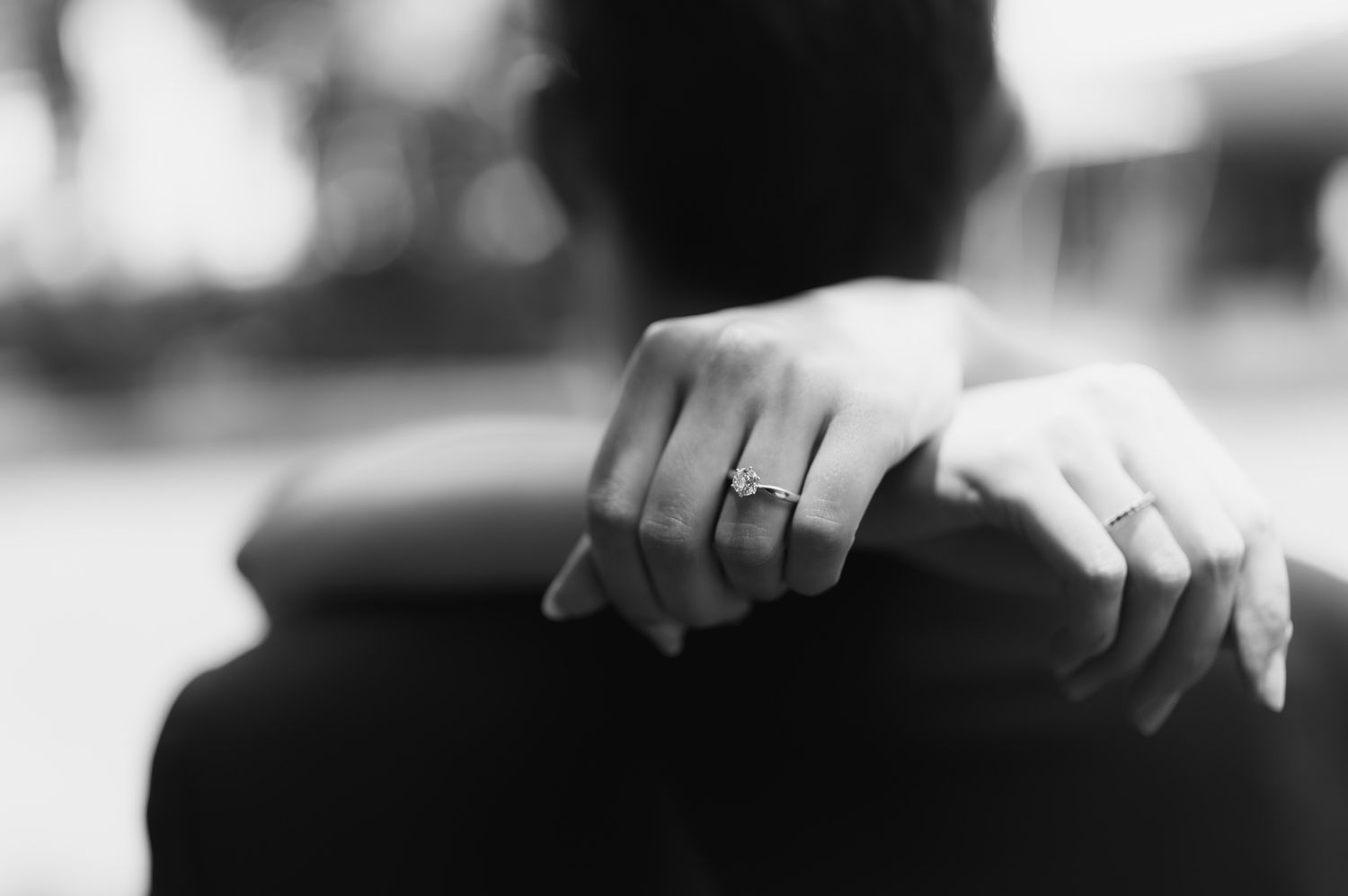 Close-up black and white photo of bride's hand with diamond engagement ring resting on groom's shoulder.