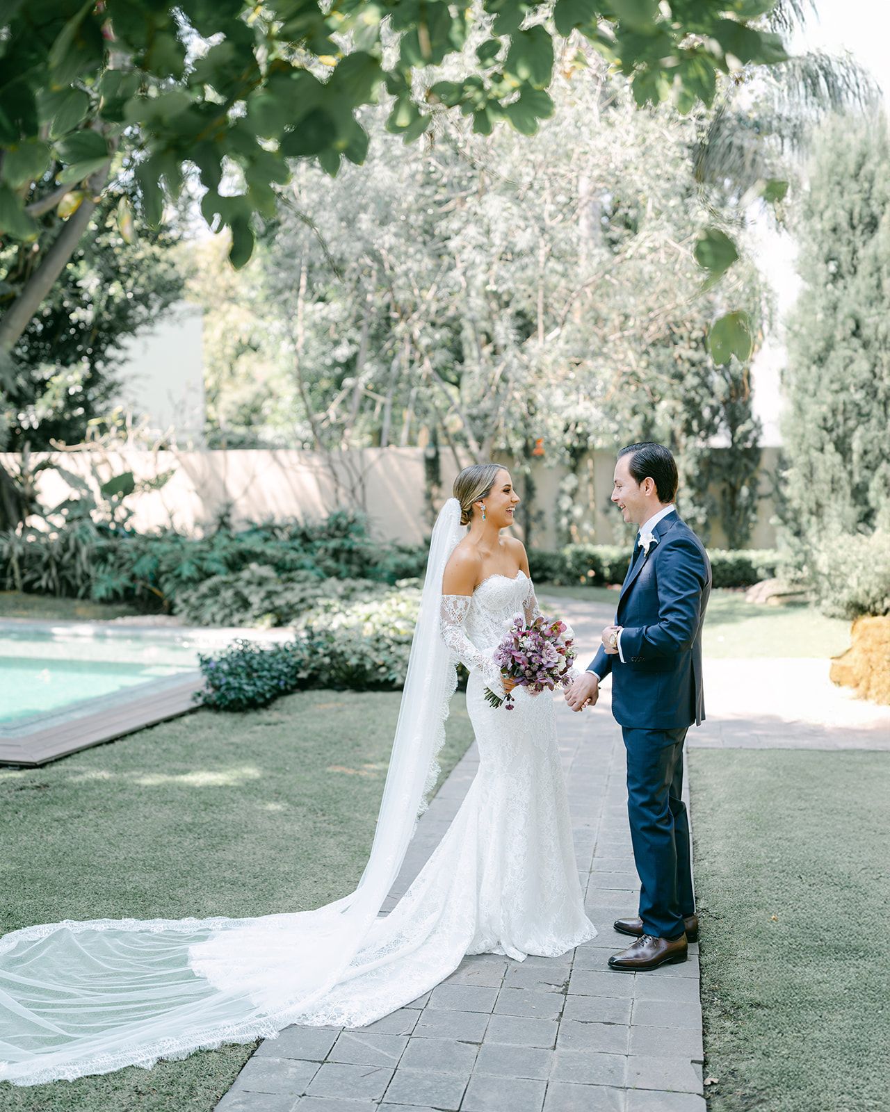 Bride and groom share a romantic first look moment in a lush garden with a pool backdrop.
