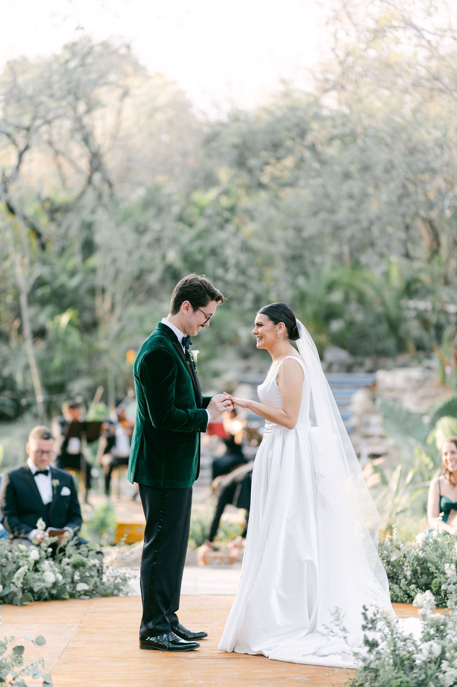 Groom placing wedding ring on brides finger during an outdoor garden ceremony with lush greenery.