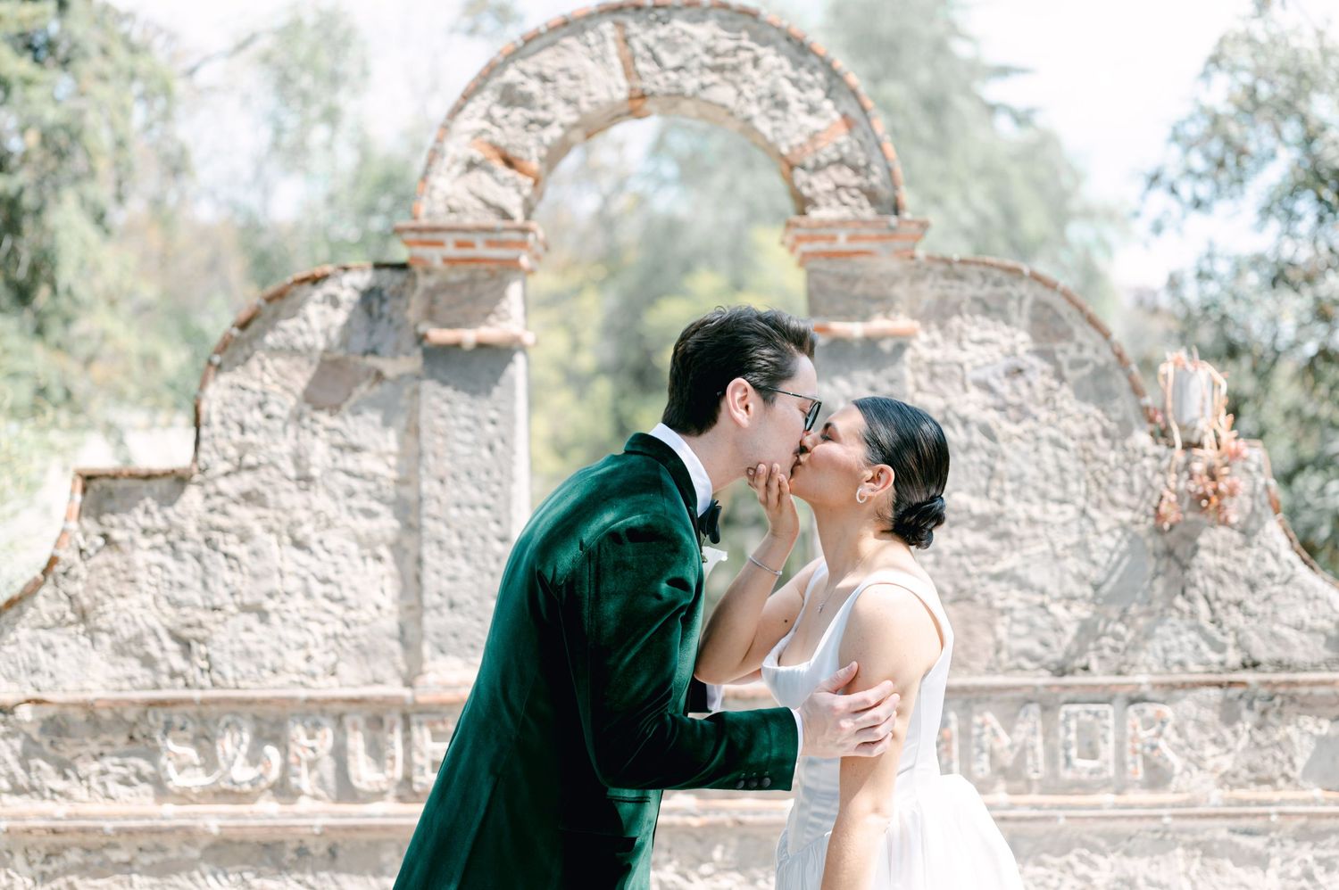 Bride and groom share a kiss in front of an ornate stone arch monument in a lush garden setting.
