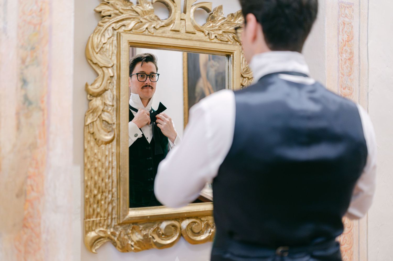 Groom adjusting his bow tie while looking into an ornate gold-framed mirror before the wedding.