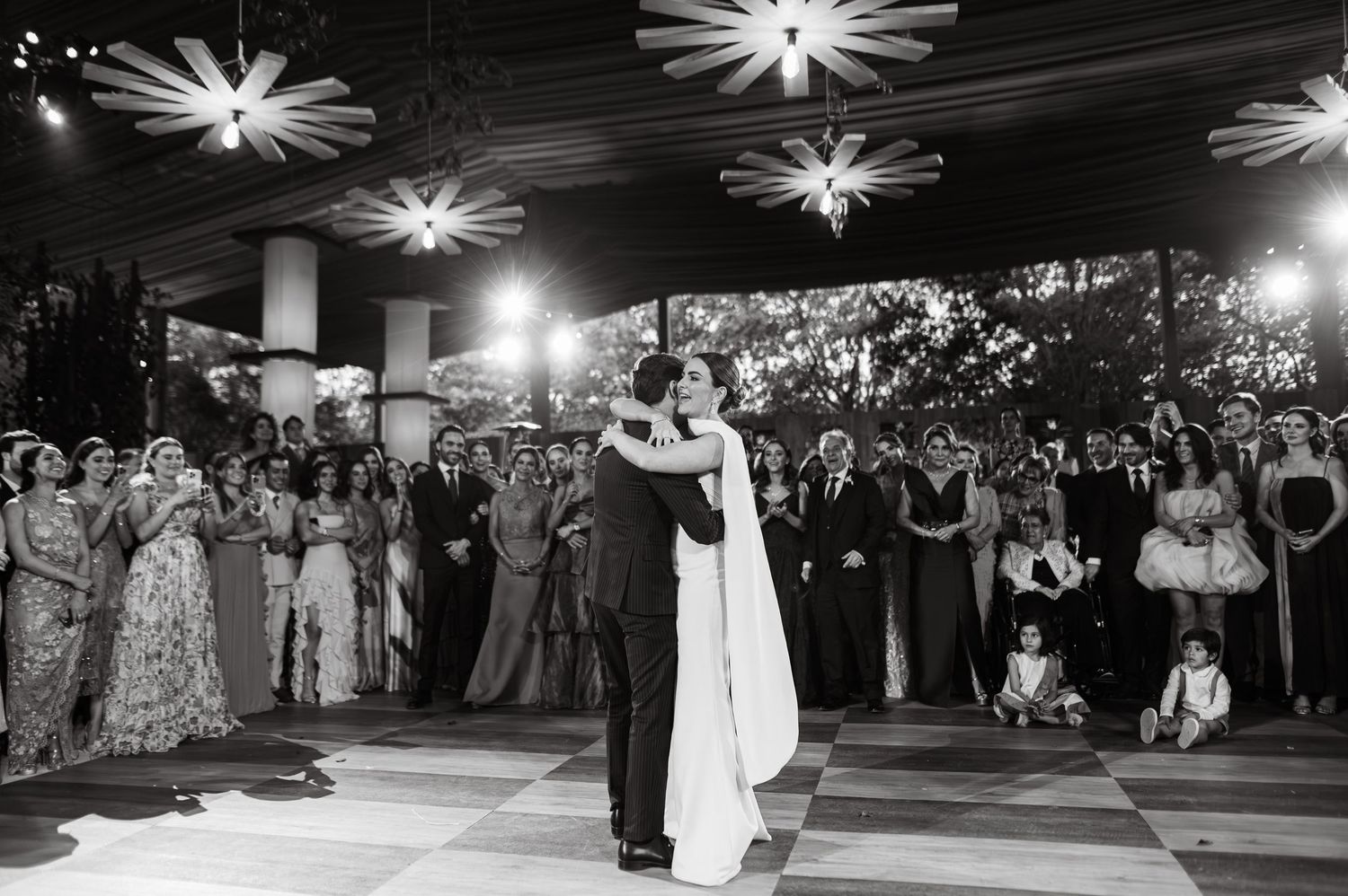 Black and white photo of bride and groom sharing first dance surrounded by wedding guests.