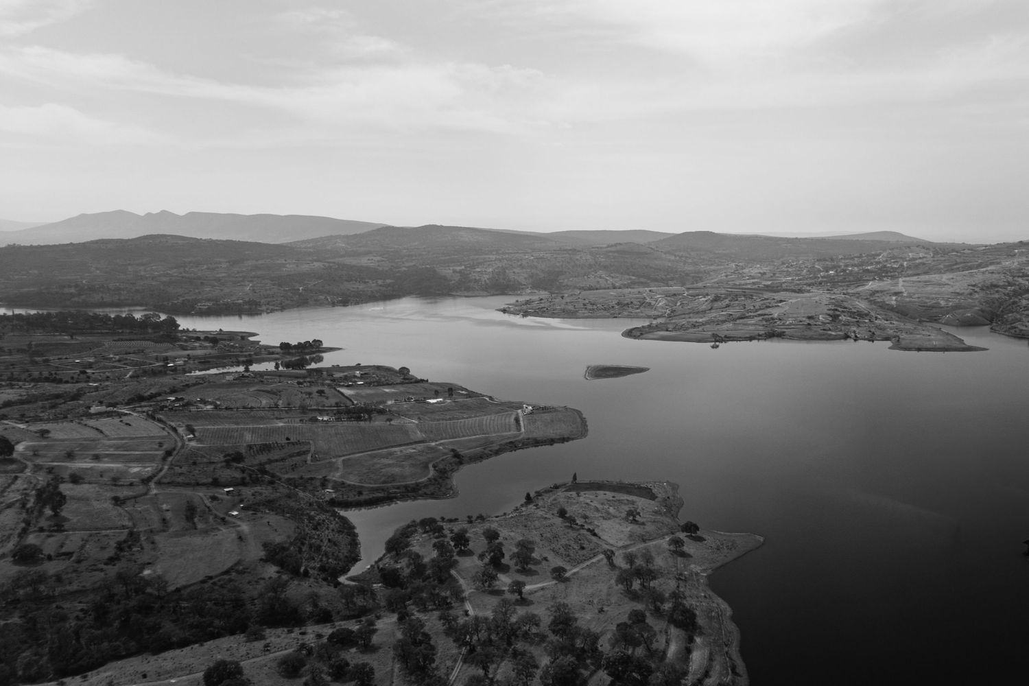 Aerial black and white landscape view of serene lake surrounded by hills and farmland.