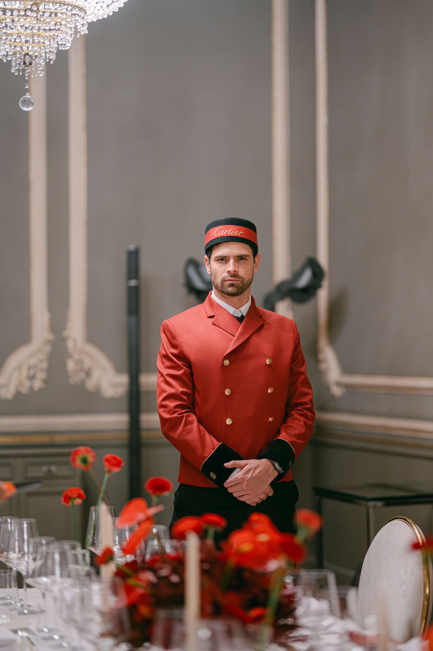 Hotel bellboy in red uniform and cap standing at attention beside a formal dining table.