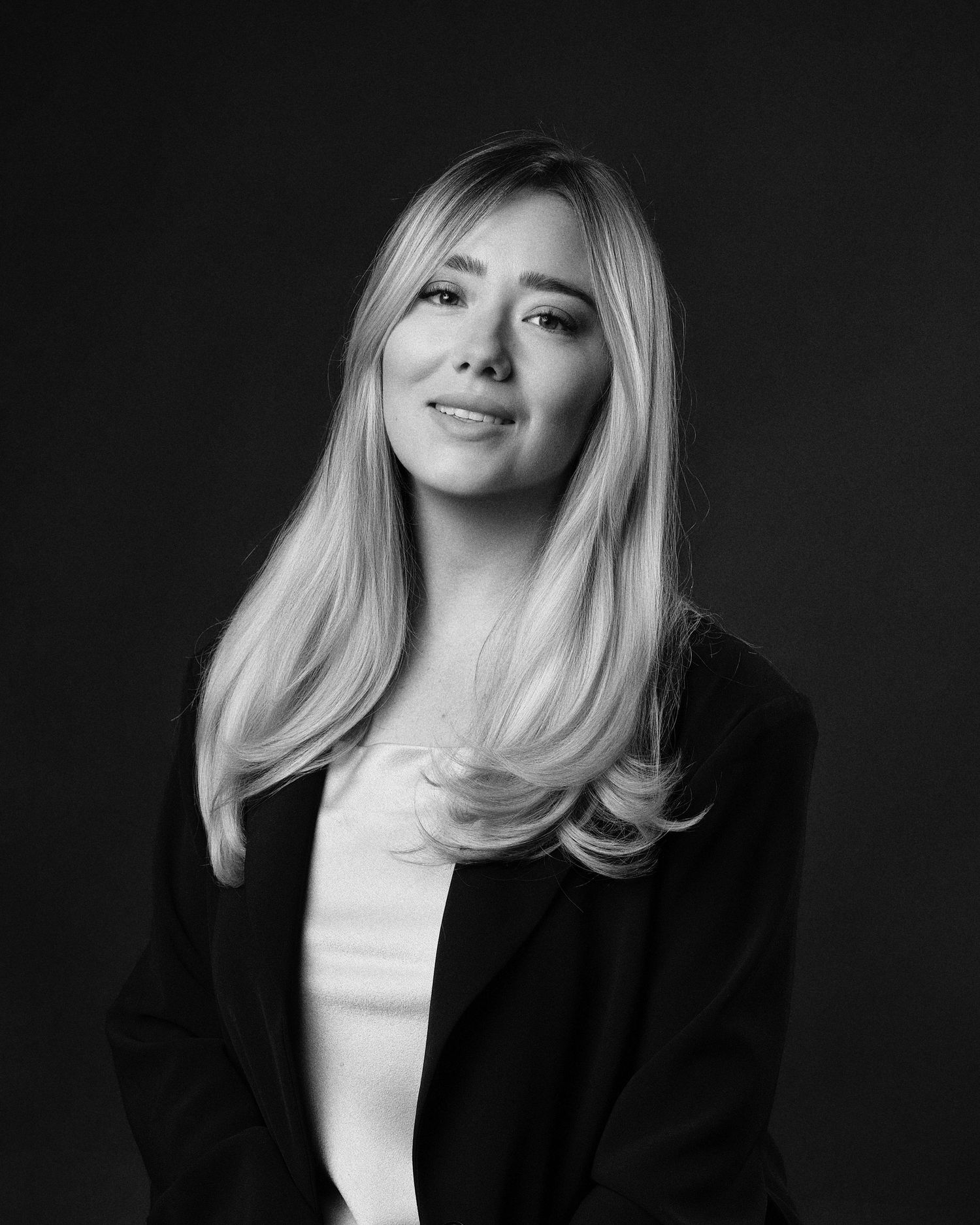 Black and white portrait of a blonde woman in a blazer smiling confidently against a dark background.