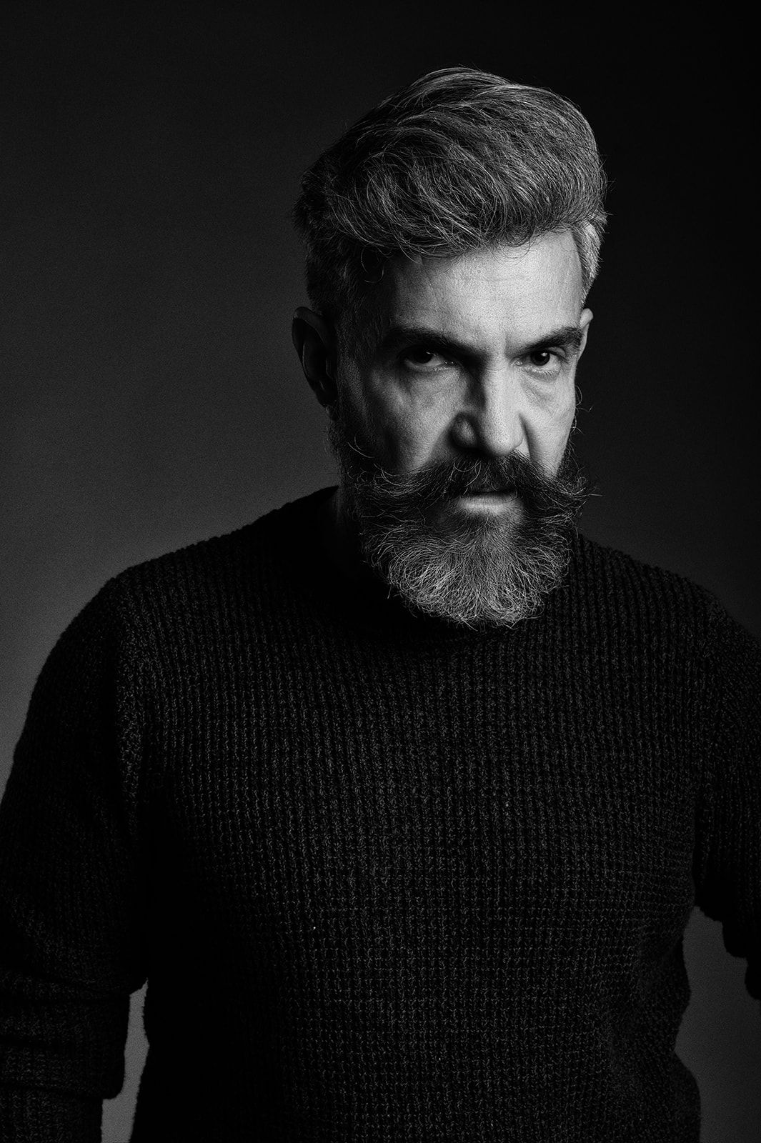 Black and white studio portrait of a serious bearded middle-aged man in a dark knit sweater.