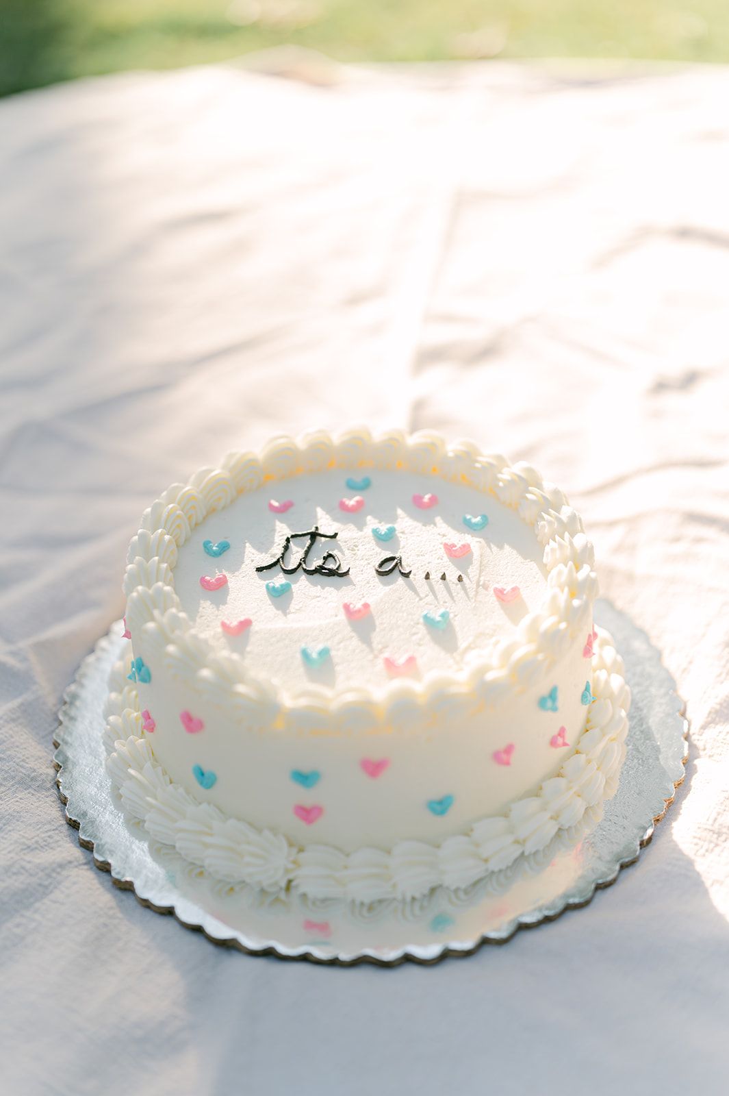 Round white gender reveal cake with pink and blue hearts and 'It's a...' text on white blanket.