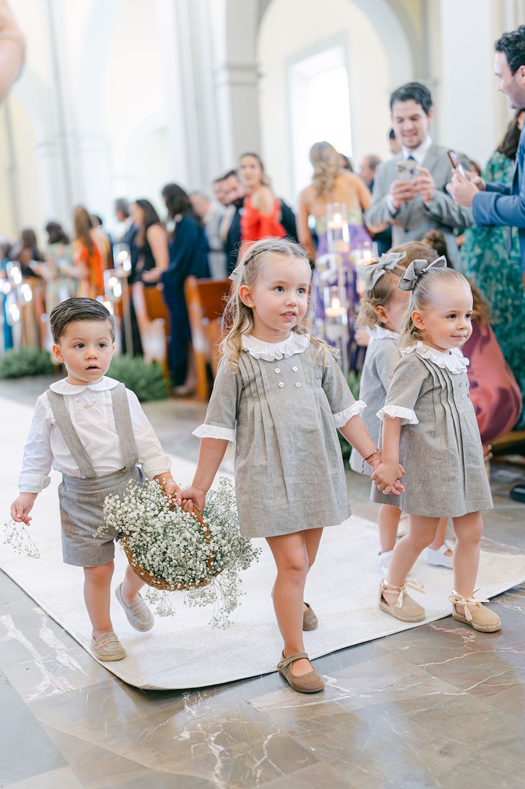 Four children in matching gray outfits walk down a church wedding aisle, with a flower girl carrying a baby's breath basket.
