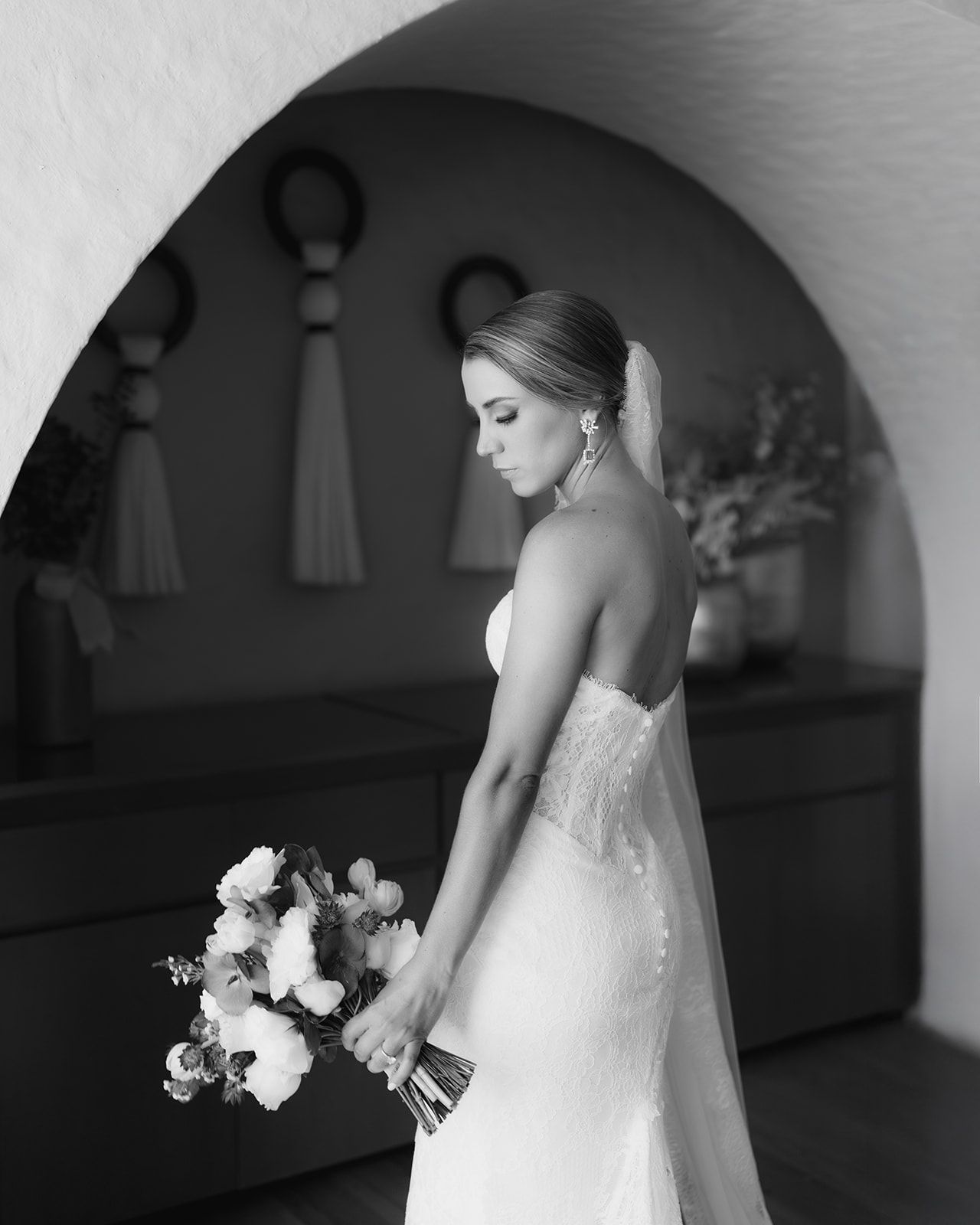 Black and white bridal portrait of a bride in lace gown holding a bouquet under an arch.