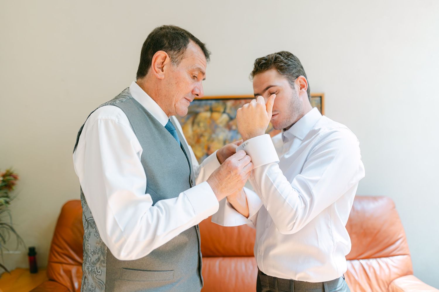 An older man helps a younger groom fasten his cufflinks while getting ready for a wedding.