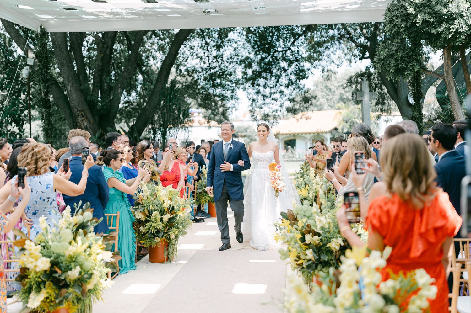 Wedding guests gather outdoors under trees with floral arrangements and decorative elements.