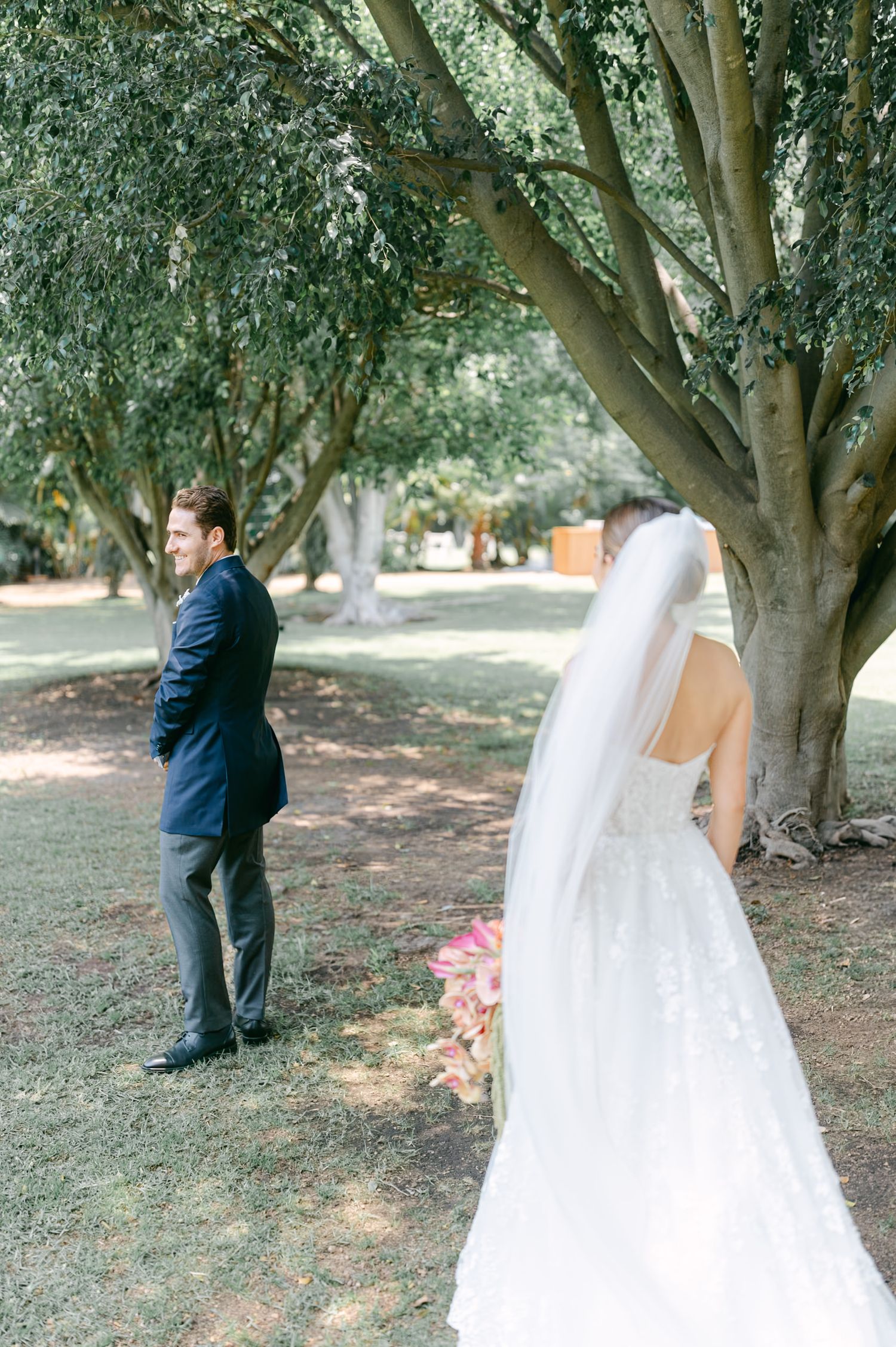Groom in navy suit standing with back turned awaiting first look with bride in lace gown.