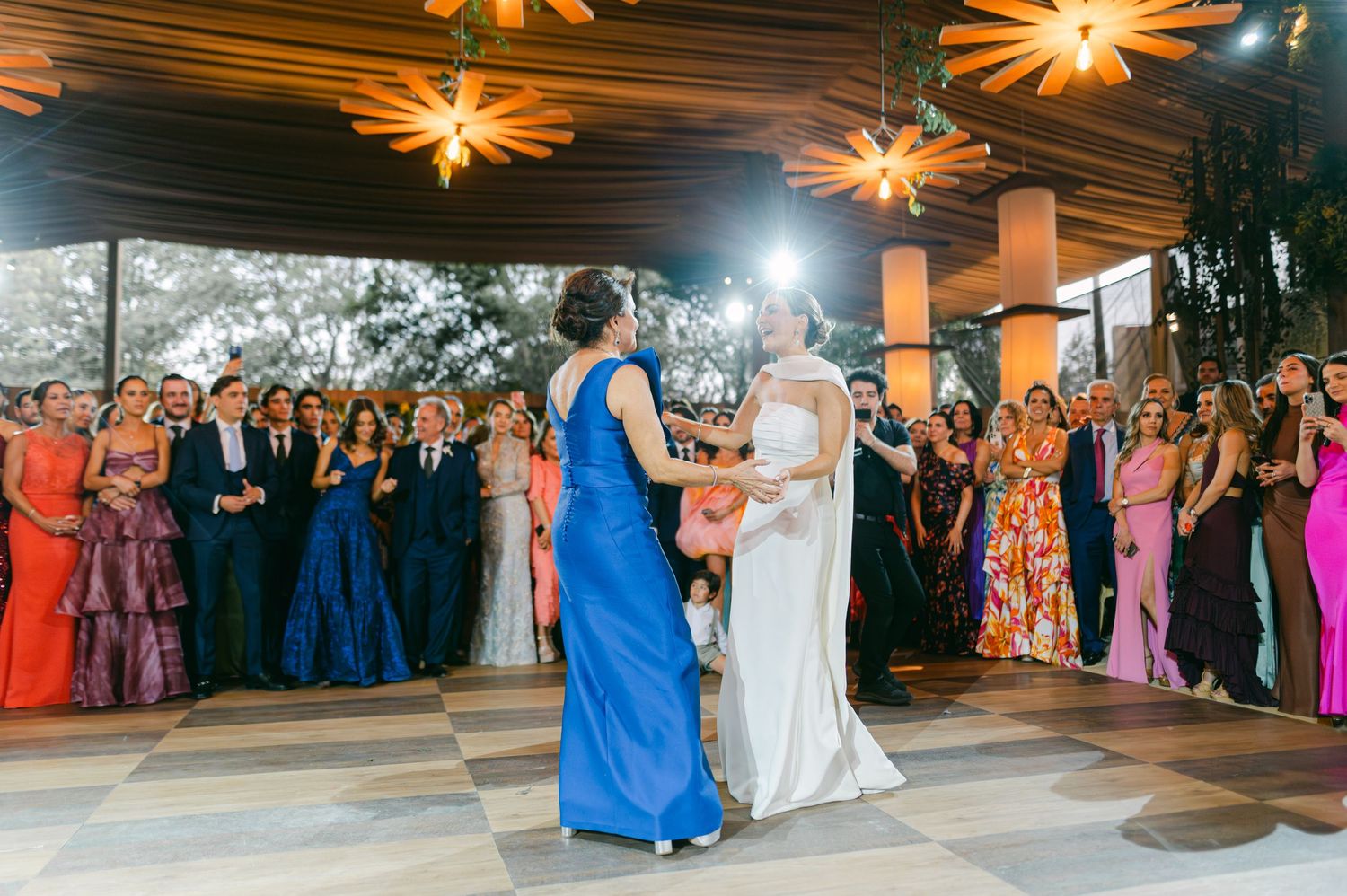 Bride in white gown dances with woman in blue dress while guests watch at elegant wedding reception.