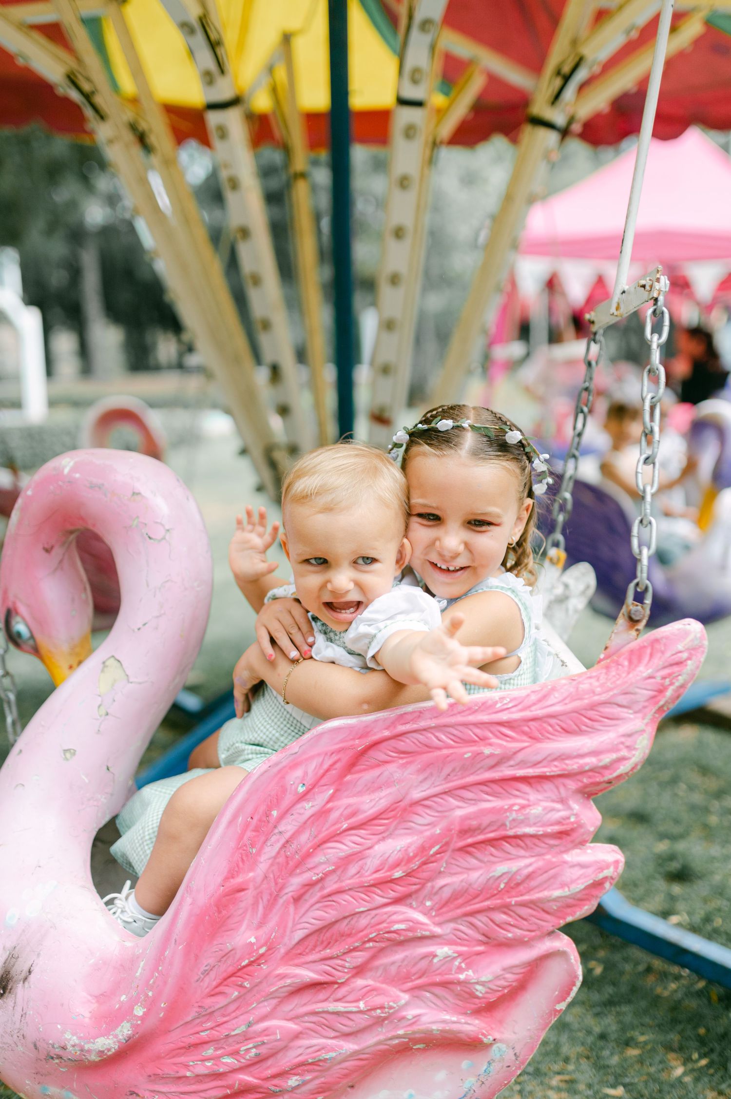 Two young children laughing and hugging on a pink flamingo carousel ride at a fair.