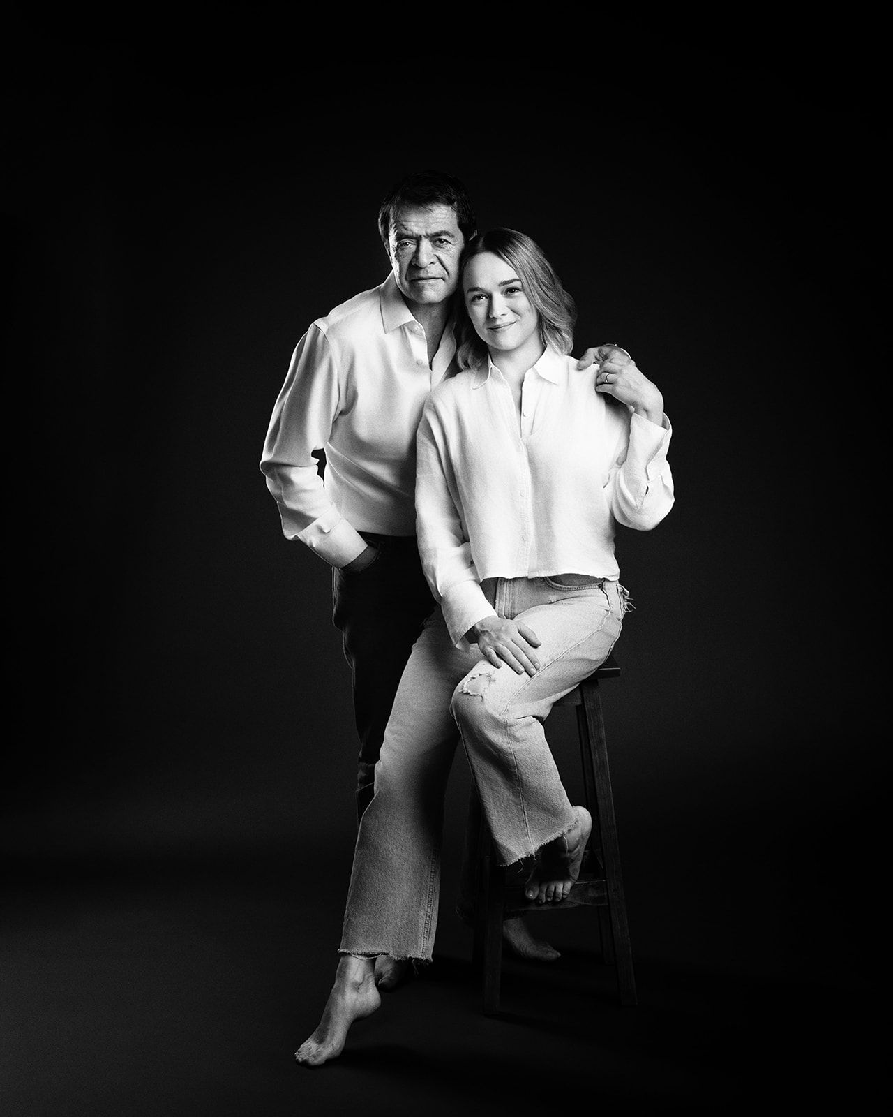 Black and white studio portrait of father and daughter posing together on dark background.