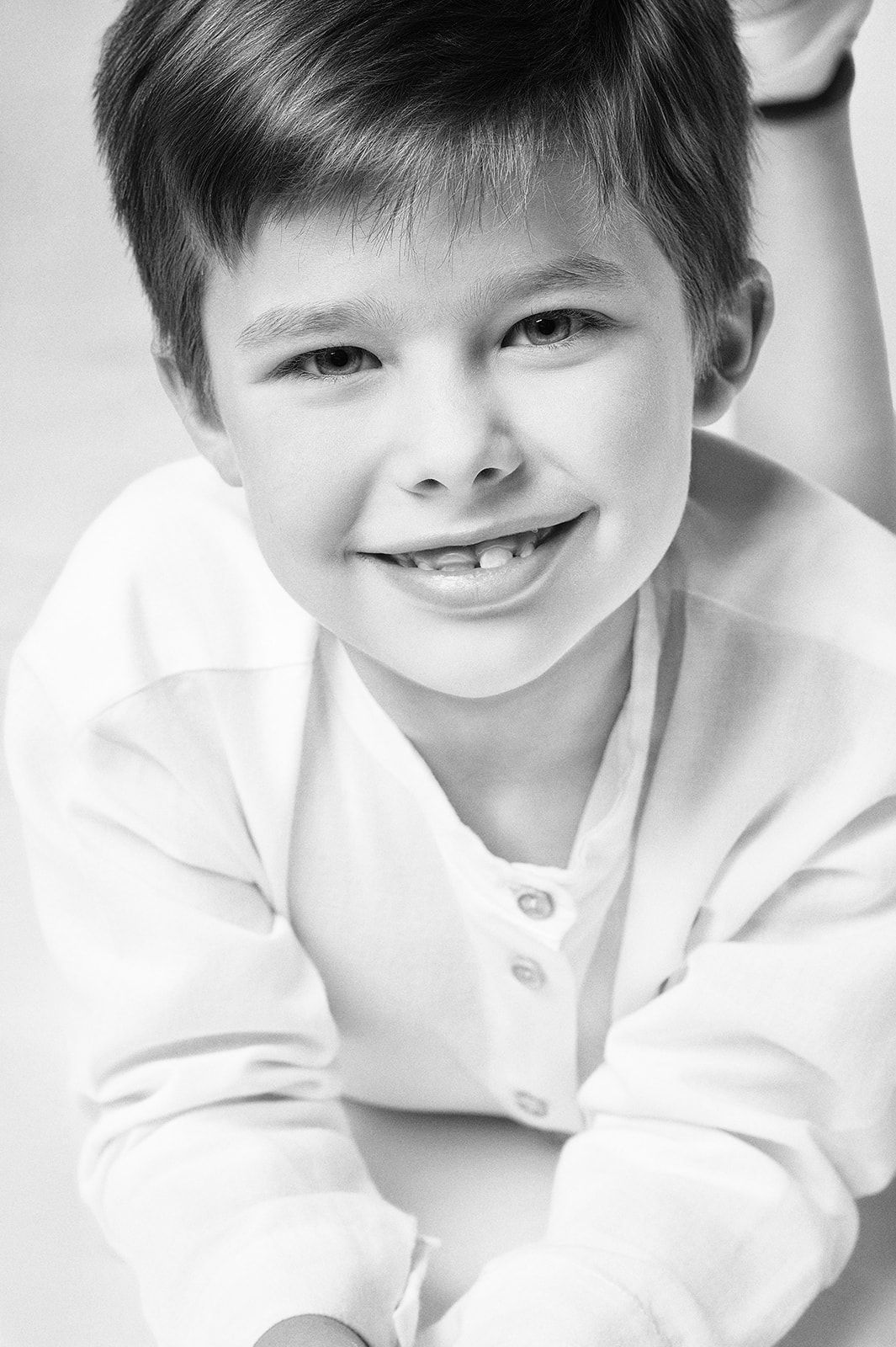 Smiling young boy in white shirt lies down looking up at camera in black and white studio portrait.