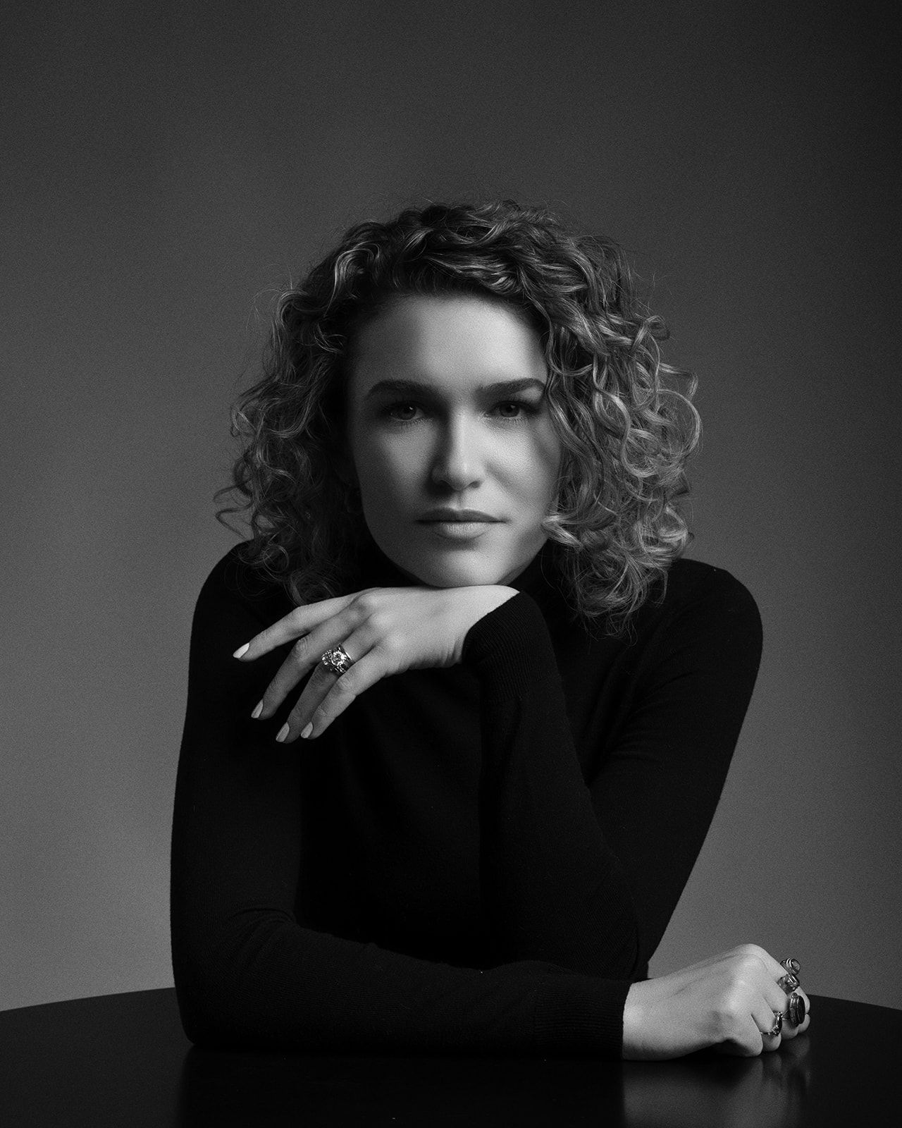 Black and white portrait of a curly-haired woman in black turtleneck resting arms on a table.