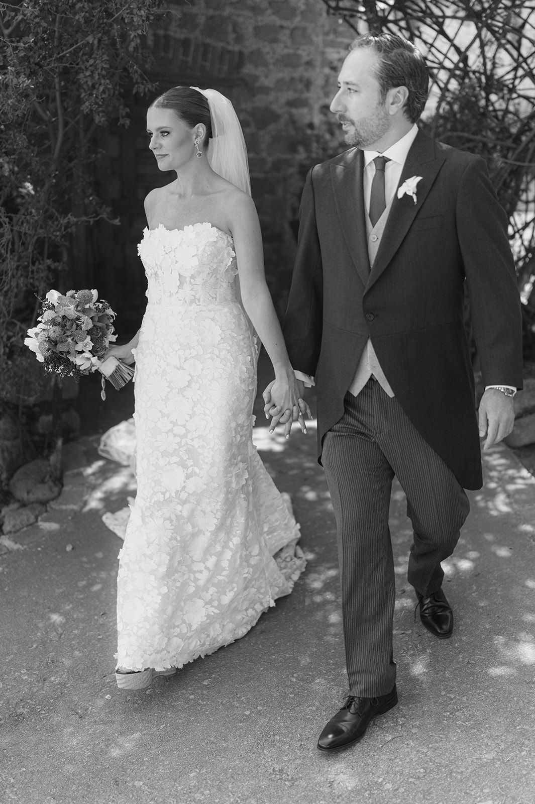 Black and white photo of a bride in a strapless lace gown holding bouquet walking with groom in suit on wedding day.
