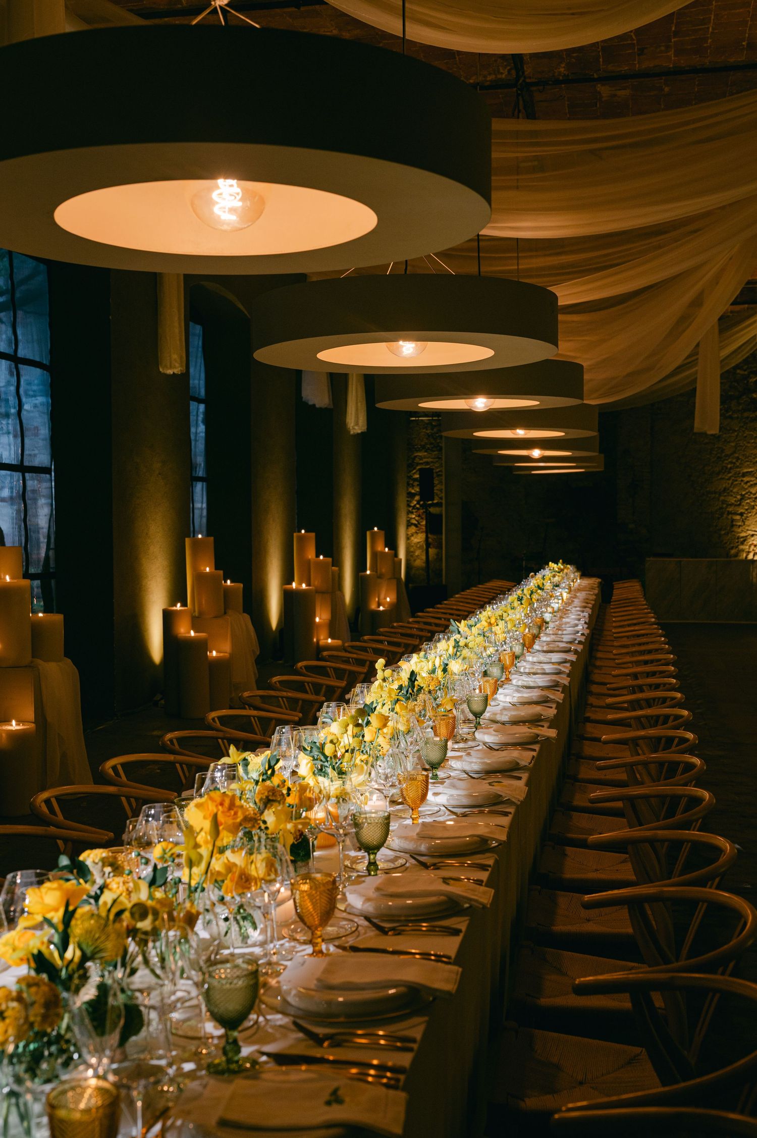 An elegantly set long dining table with yellow floral centerpieces and candlelight.