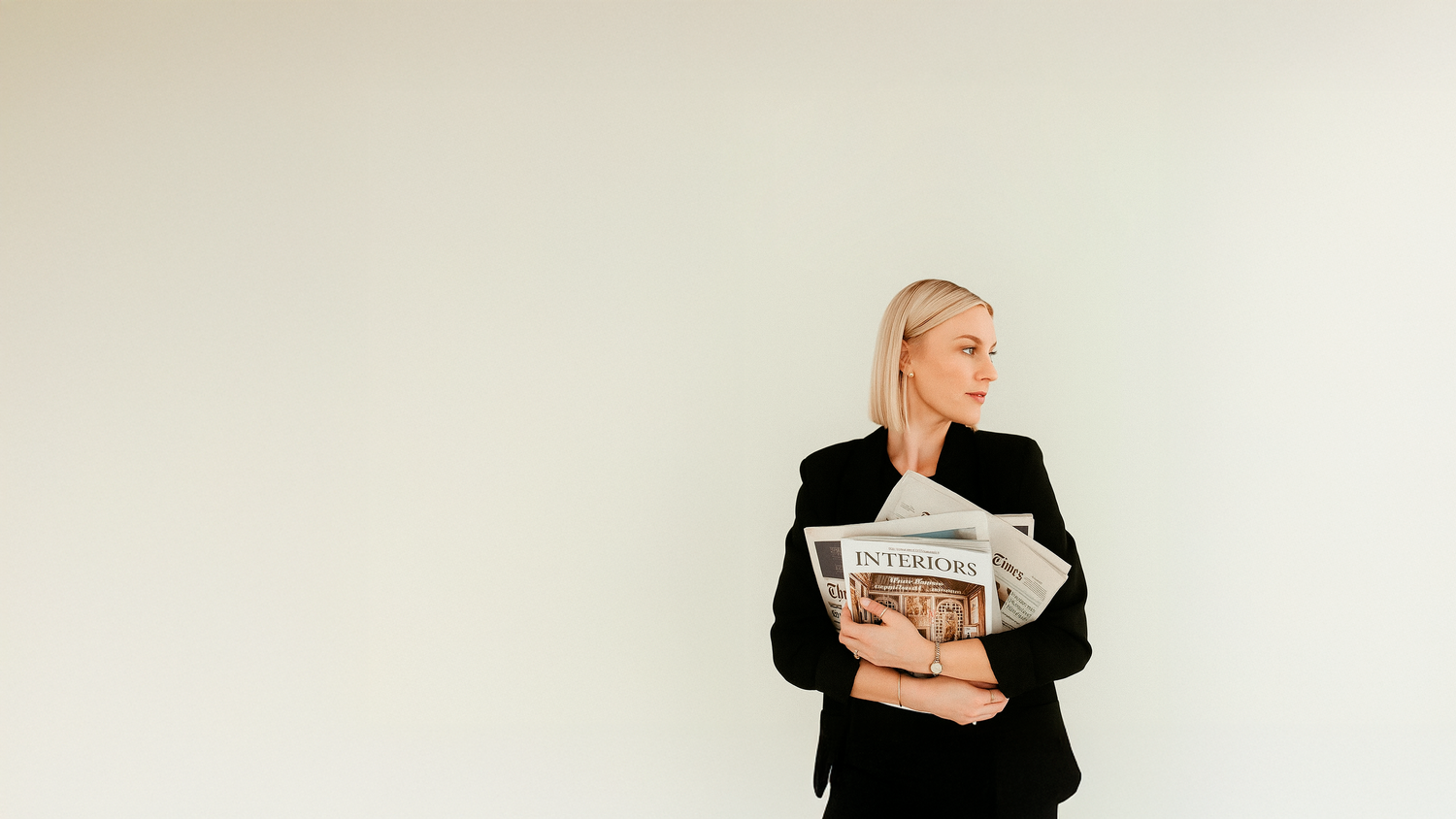 Blonde woman in black blazer holding newspapers against white wall.
