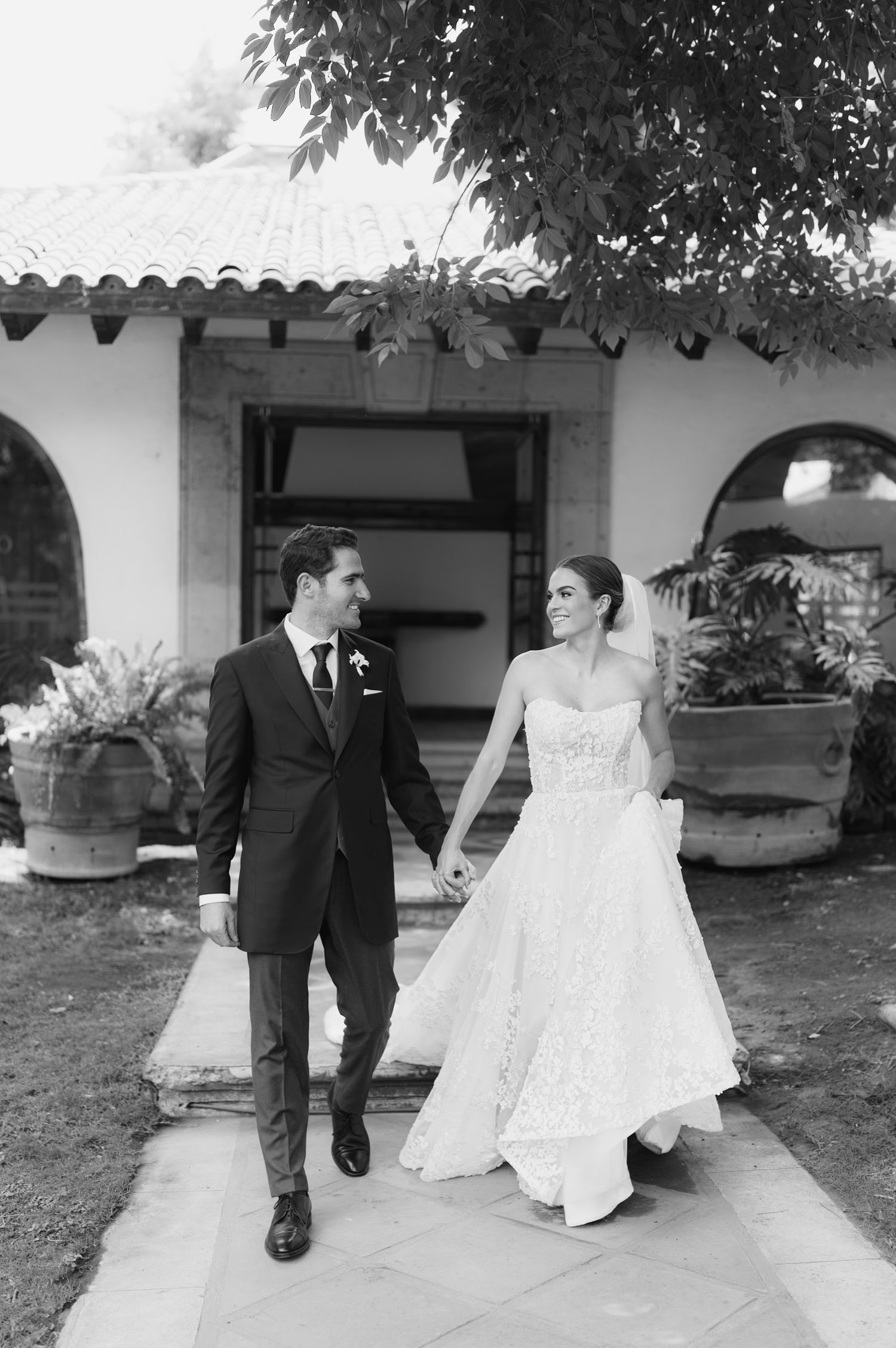 Black and white photo of smiling bride and groom walking hand in hand outside wedding venue.