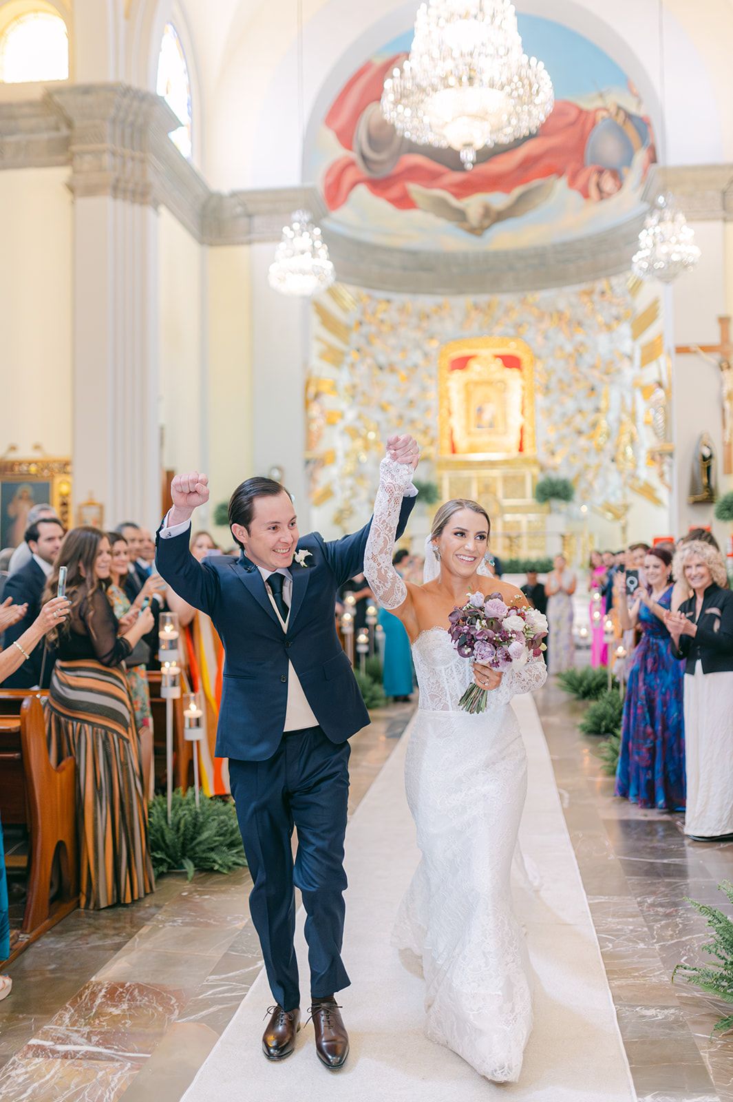 Joyful newlyweds raise hands walking down church aisle after wedding ceremony with colorful guests applauding.