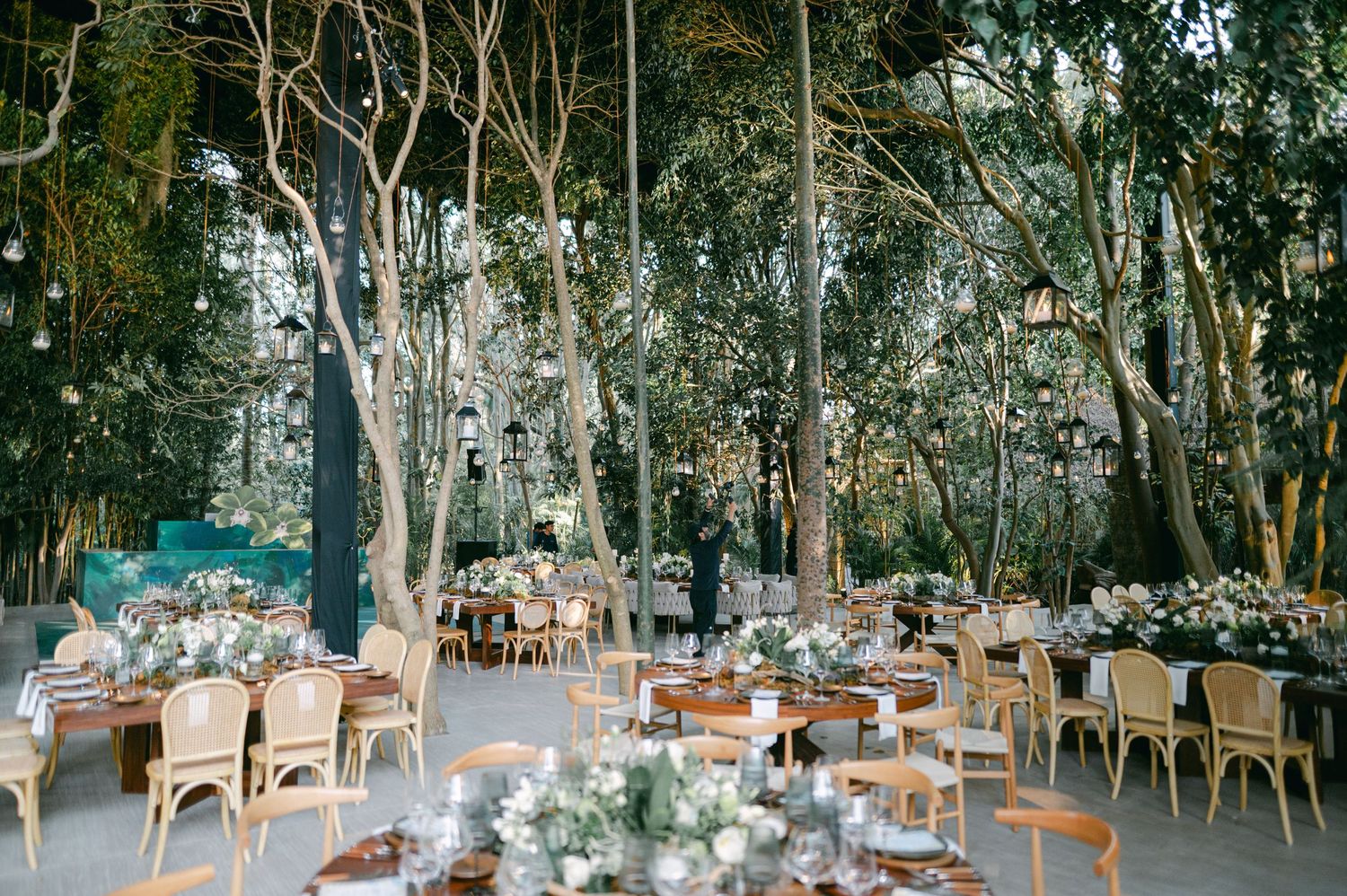 Elegant outdoor wedding reception with round tables, rattan chairs, and hanging lanterns amid lush forest trees.