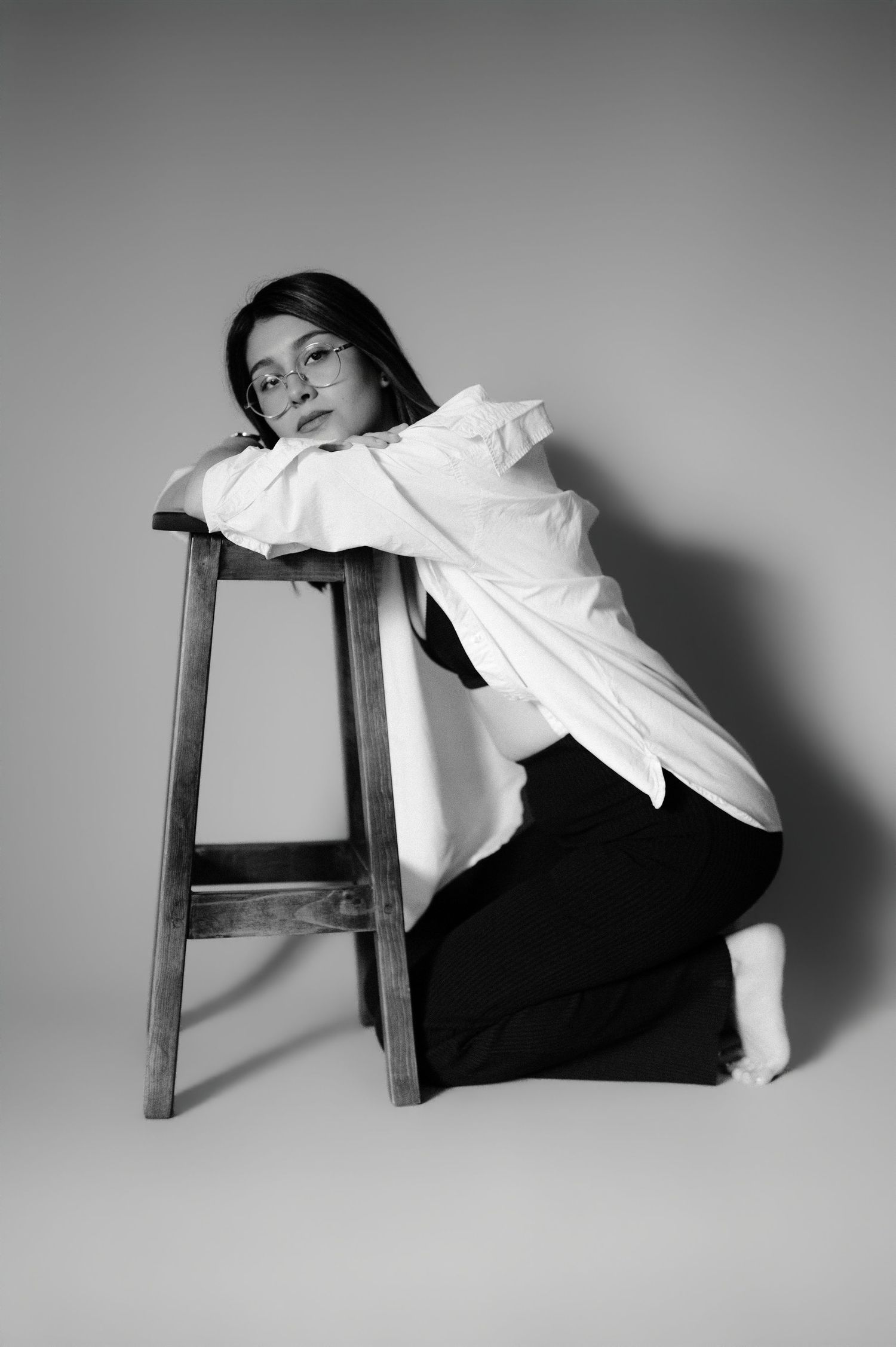 Woman in white shirt leaning on wooden stool, black and white studio portrait.