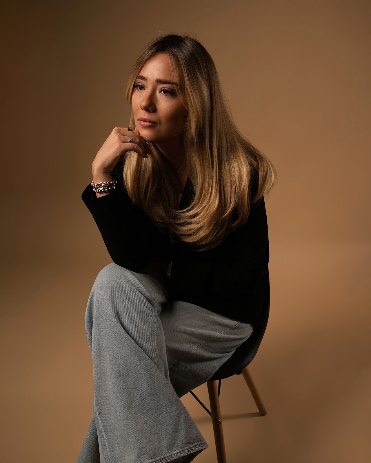 Young woman with long blonde hair sitting on a stool in a black sweater and wide-leg jeans.