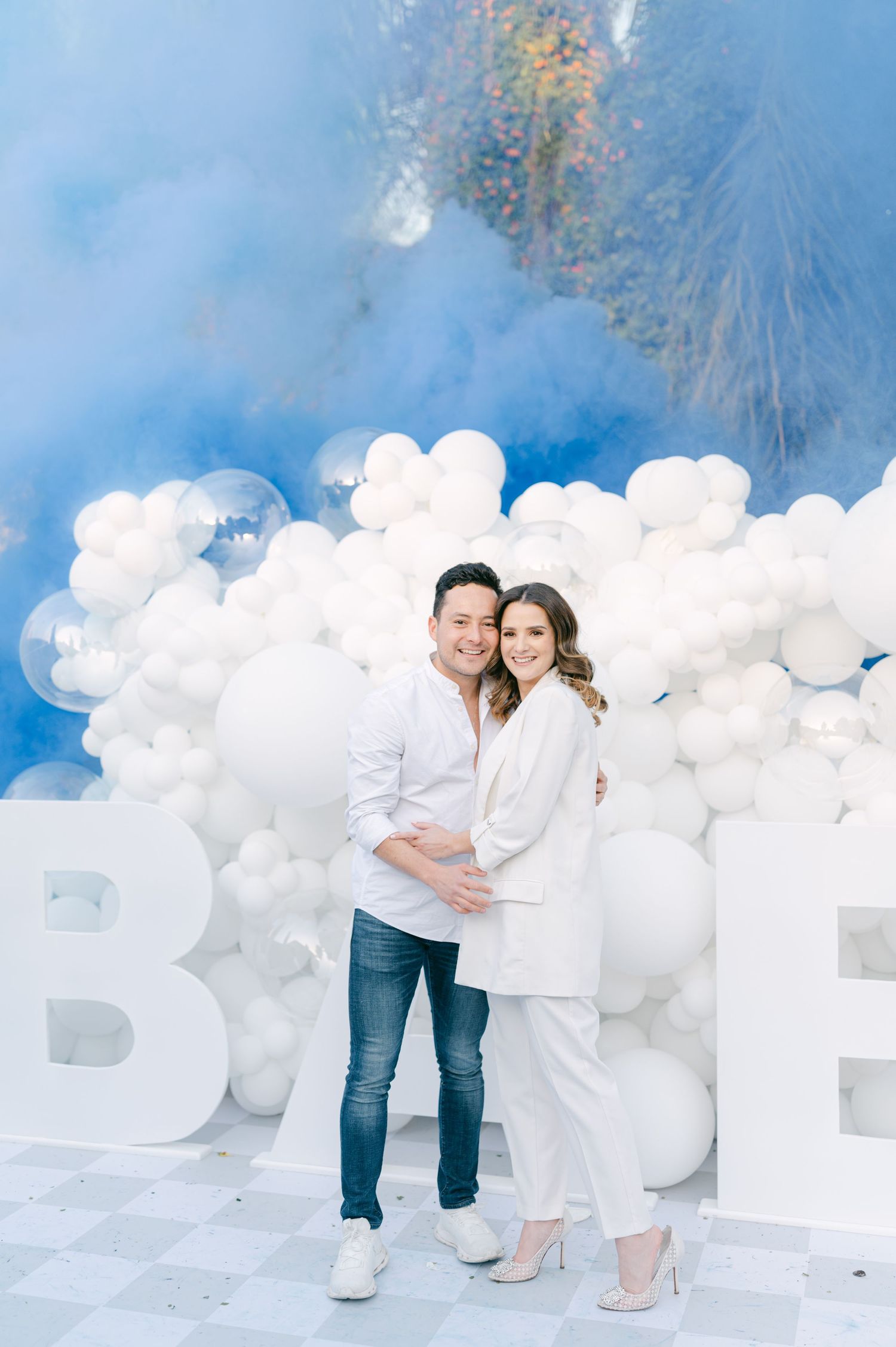 A couple embraces in front of a white balloon wall with blue smoke at a gender reveal.