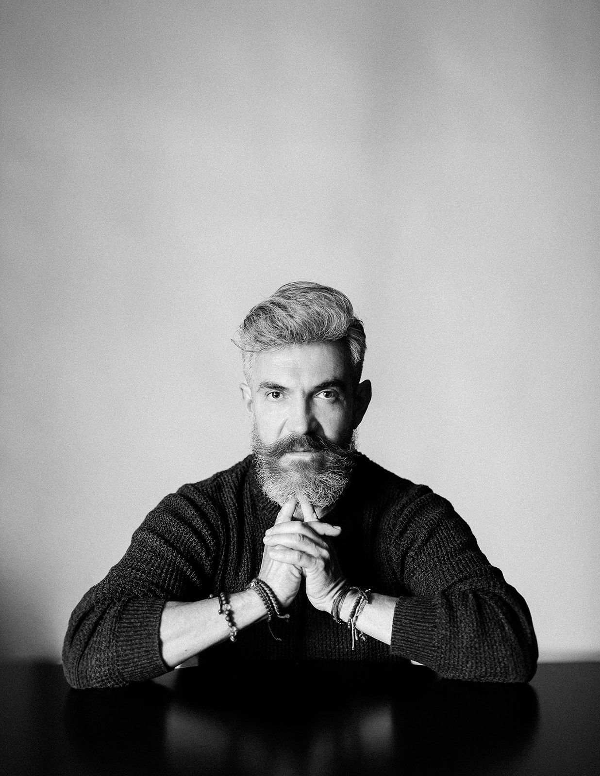 Black and white portrait of bearded man in sweater sitting at table with hands clasped.