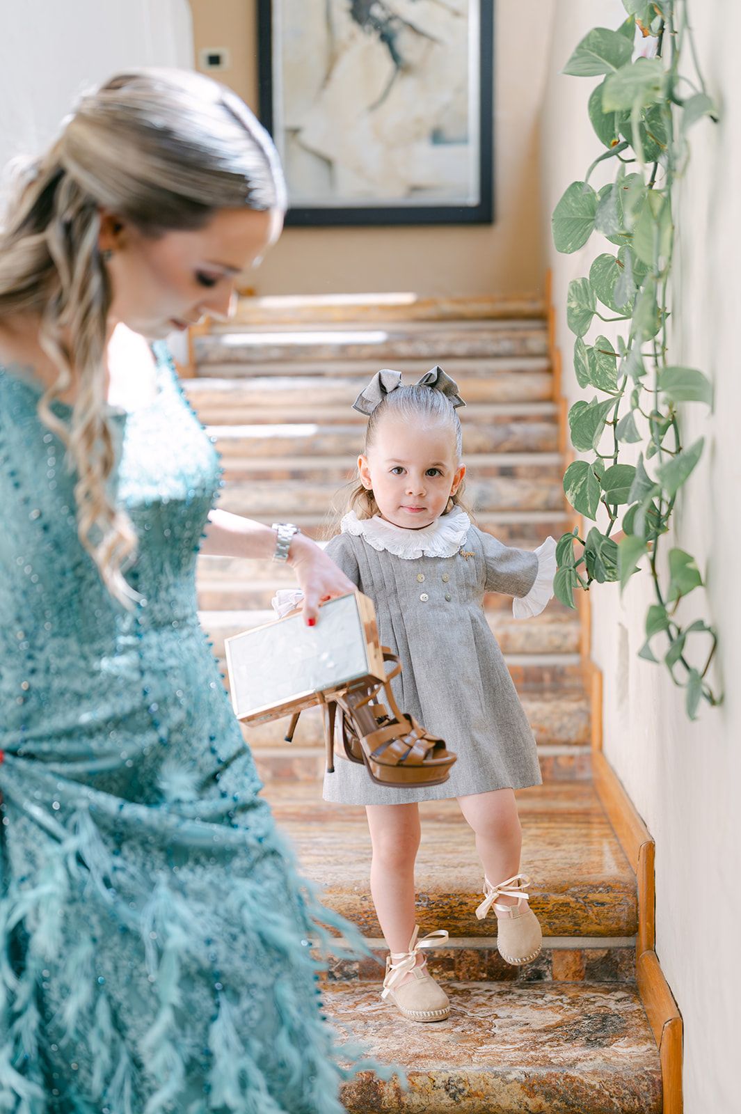 Young flower girl in gray dress holds bride's shoe on staircase decorated with greenery garland.