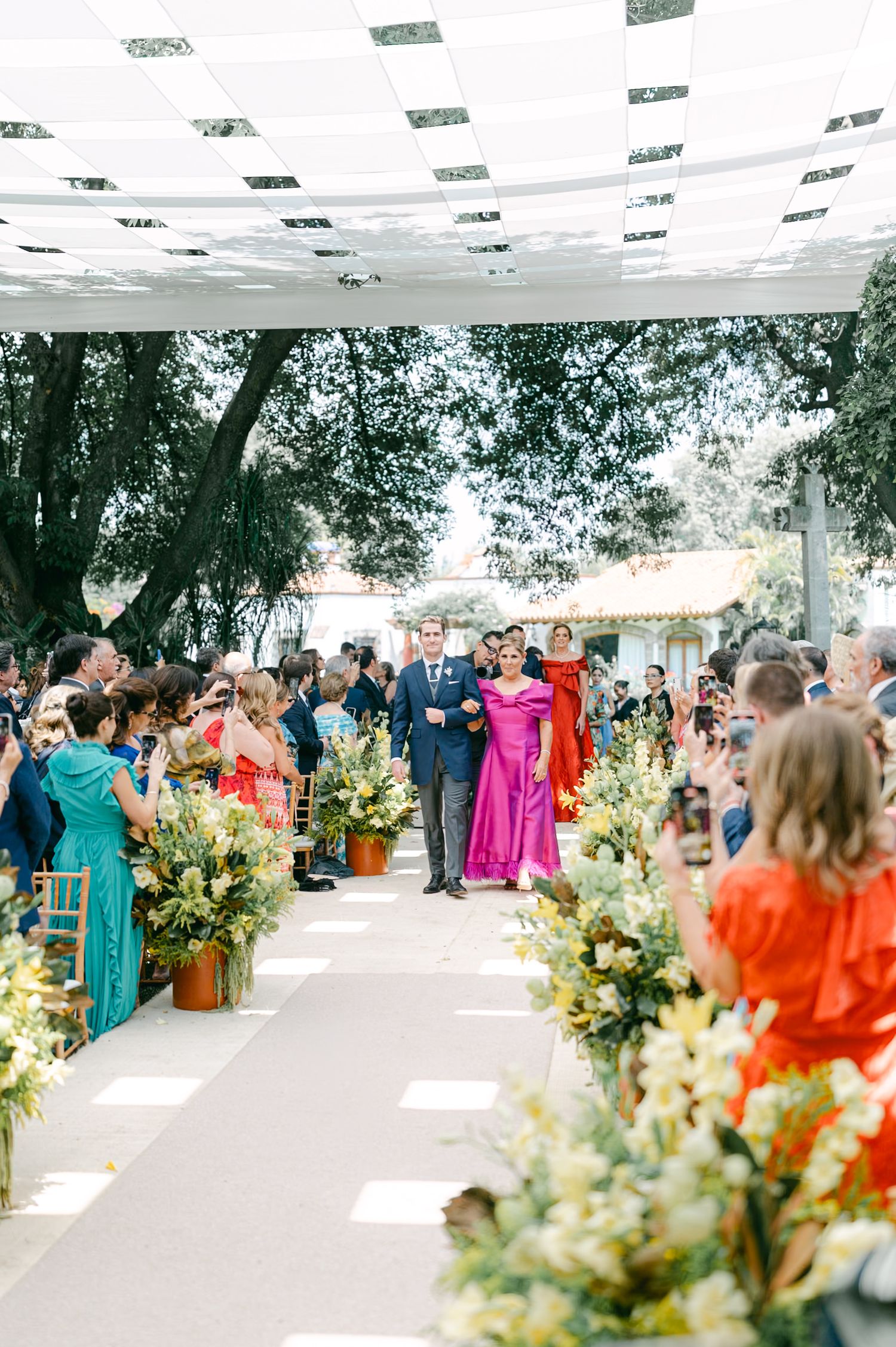 Two flower girls escorting a baby in a padded wooden cart down the outdoor wedding ceremony aisle.