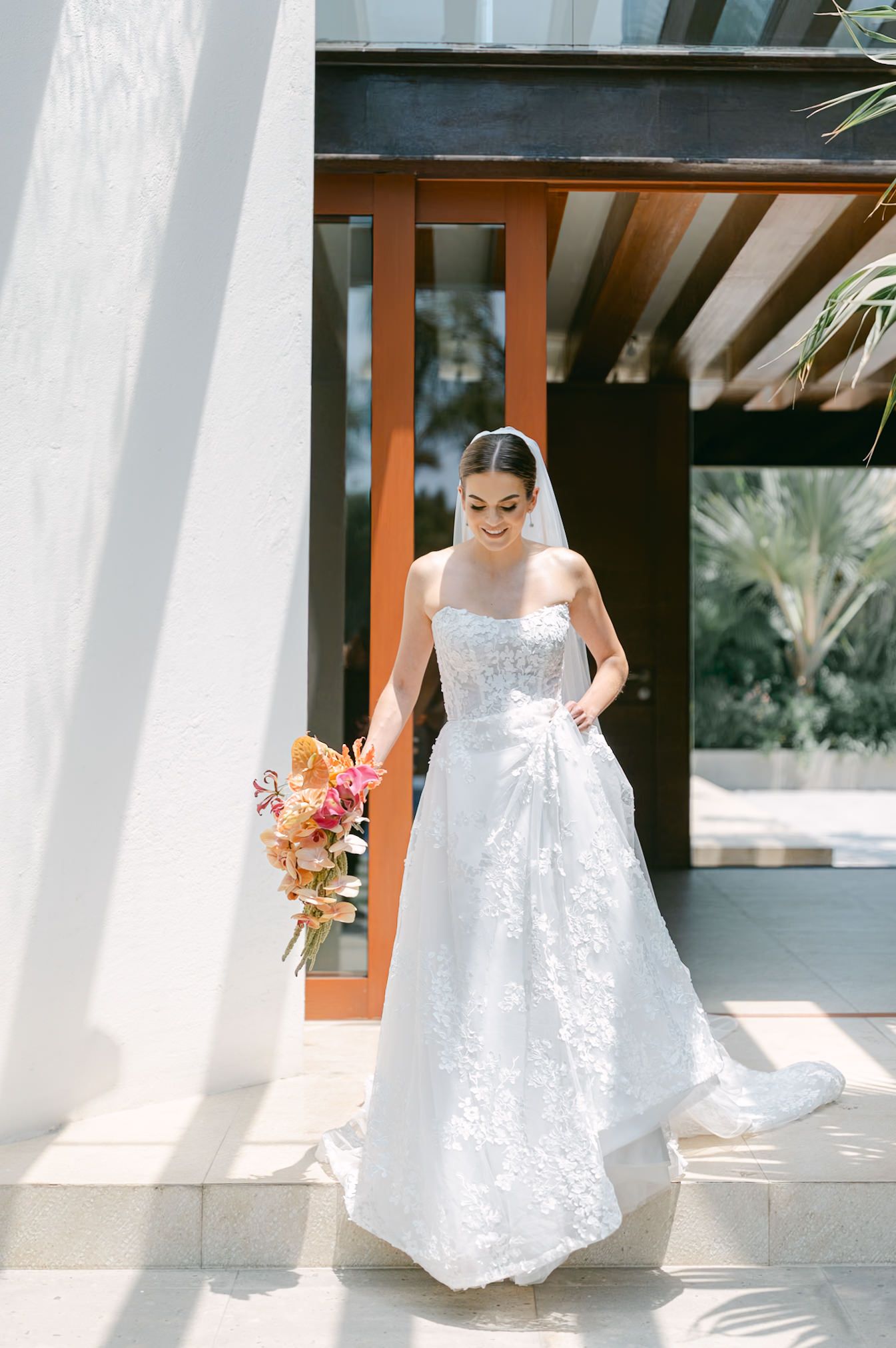 Radiant bride in strapless lace A-line gown holding vibrant pink and orange bouquet outside elegant venue.