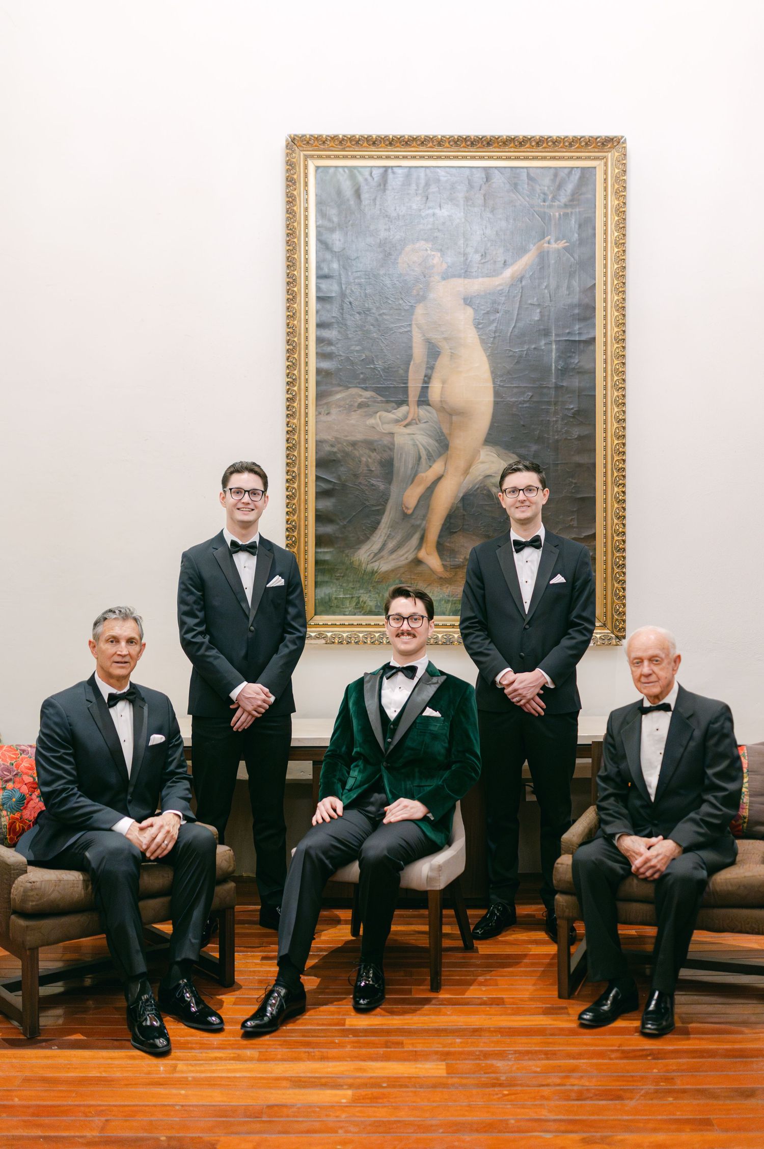 Groom in green velvet tuxedo seated with groomsmen in black tuxedos before a classical painting.