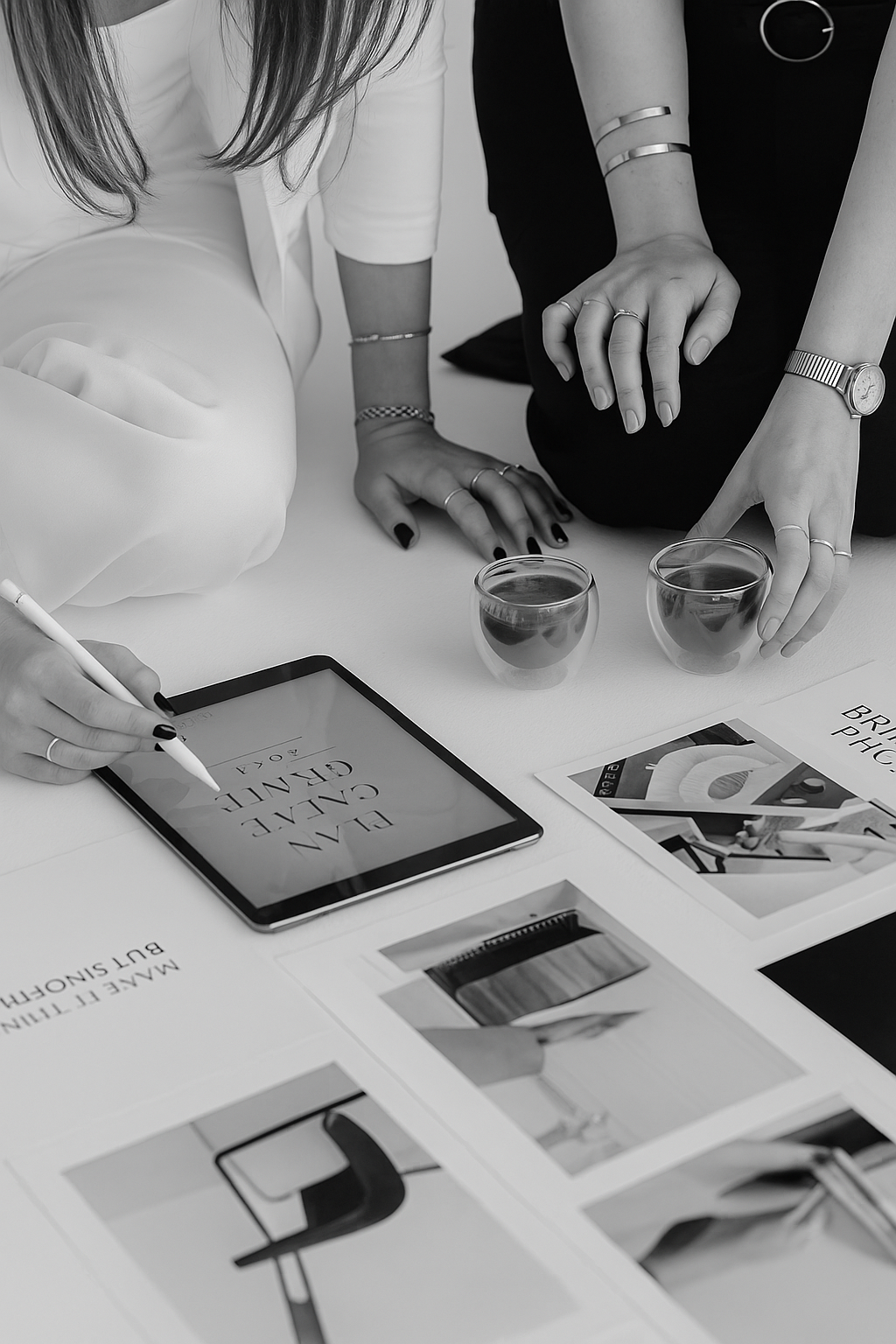 Two women reviewing design layouts on tablet and printed photos on white table.