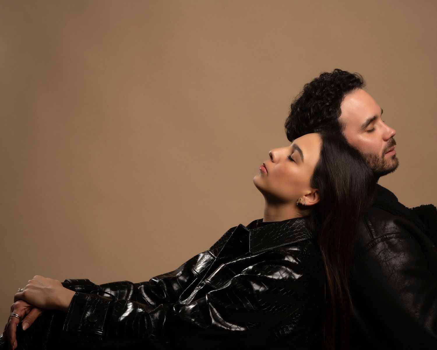 Couple in black leather jackets pose back-to-back with eyes closed in artistic studio portrait.
