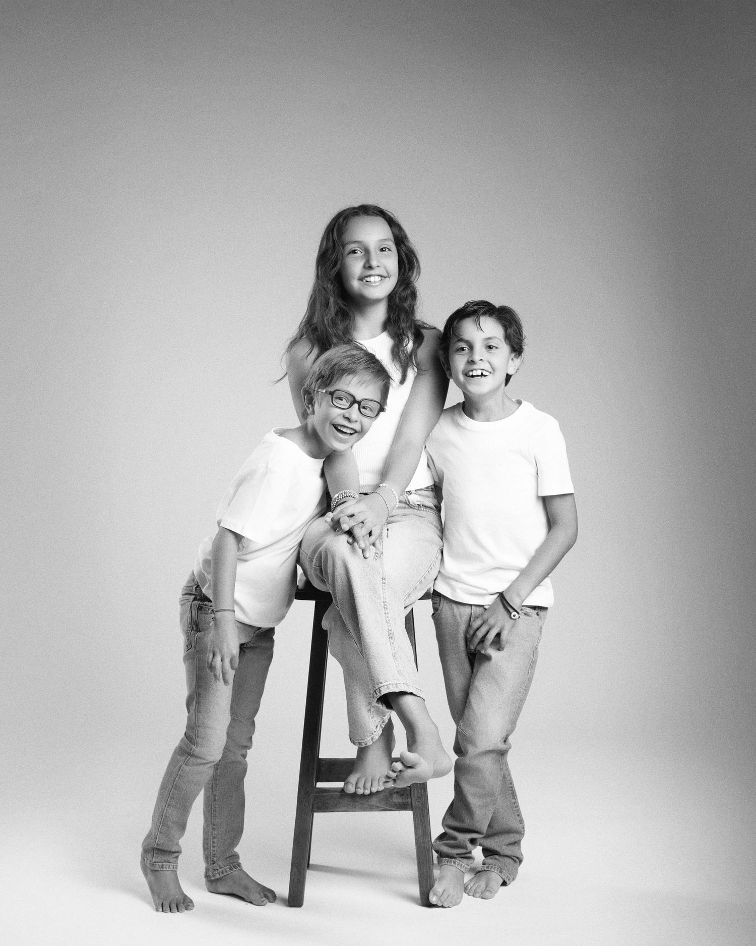 Black and white studio portrait of three children in white shirts and jeans posing on a wooden stool.