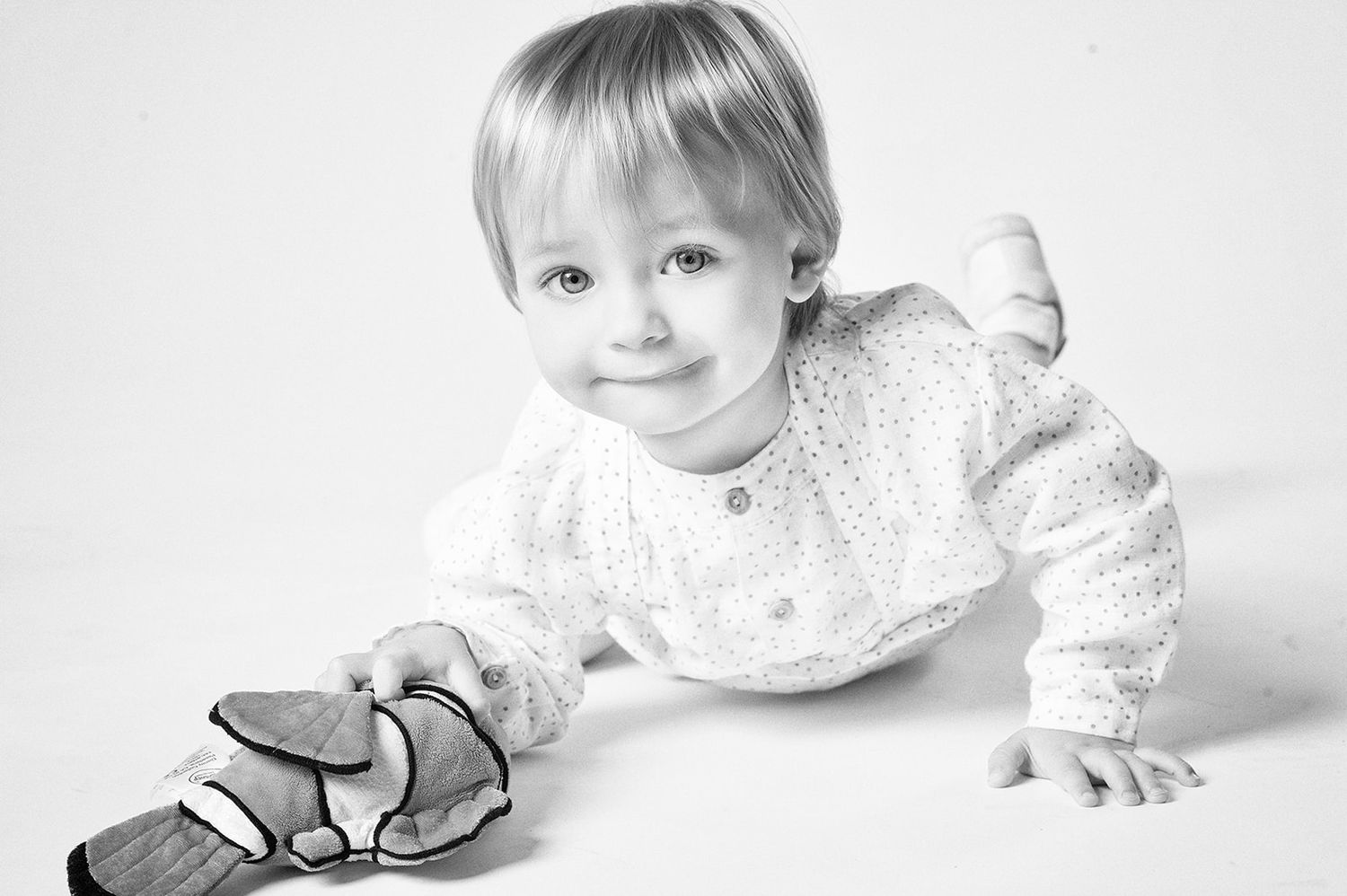 Adorable blonde toddler in white lace outfit crawls on floor holding a shoe in black and white photo.