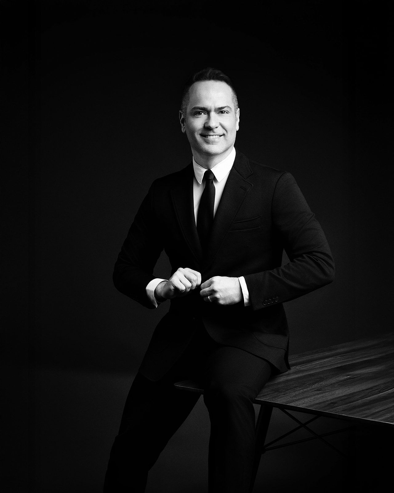 Man in a black suit and tie smiling while leaning against a table in a dark studio.