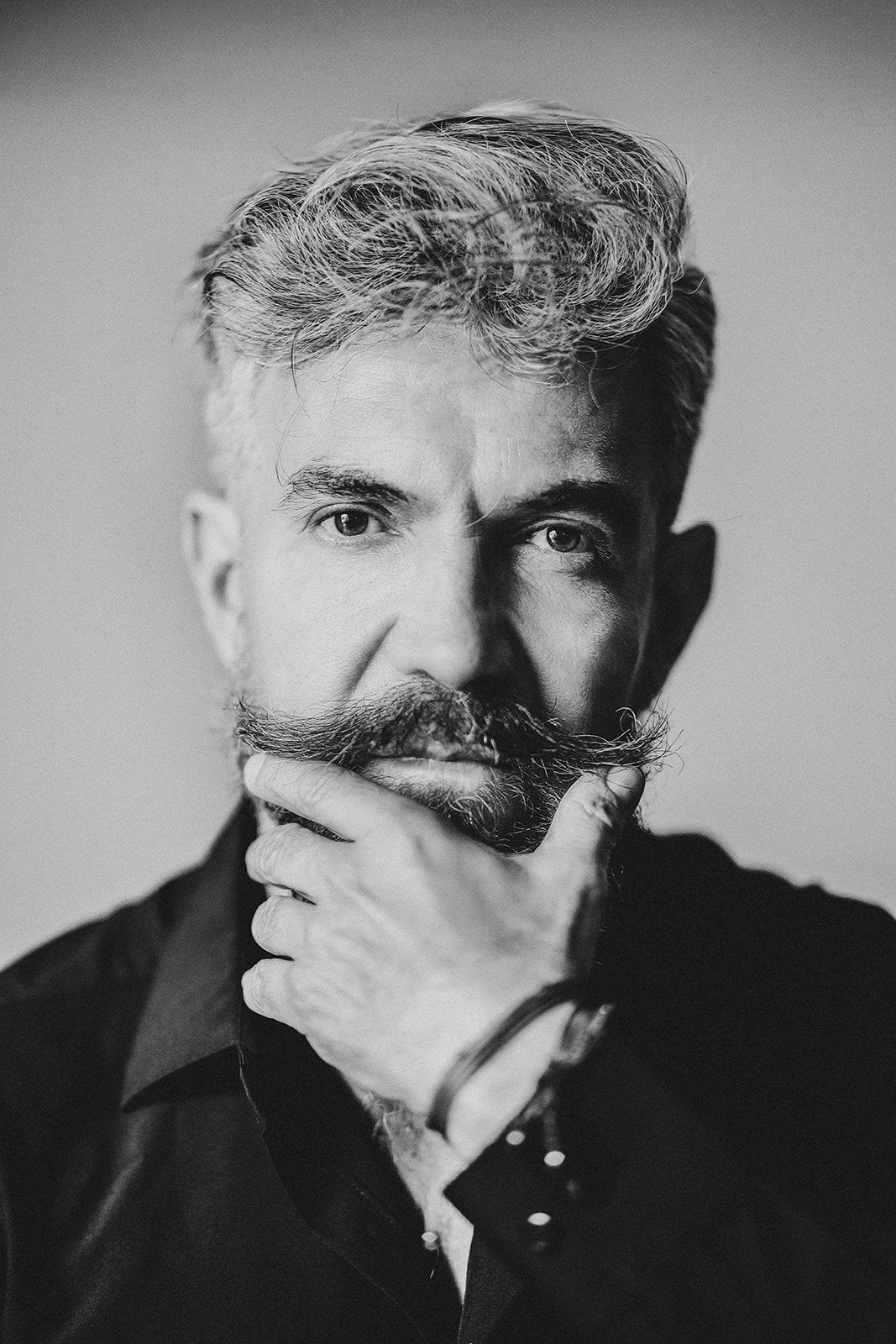 Black and white portrait of a bearded middle-aged man with gray hair, hand on chin, wearing a dark shirt.